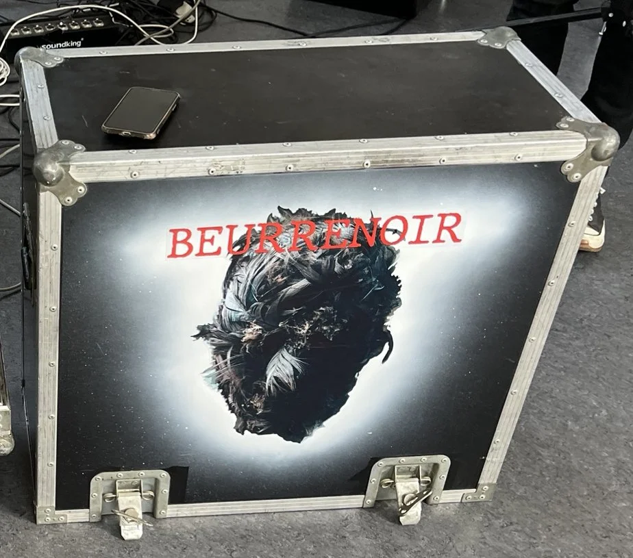 A black device case with the word "BEURRENOIR" in red text and a black and white image of a feathered bird head on the front, placed on a black carpeted floor.
