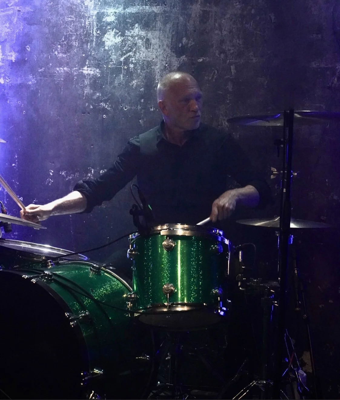 A man playing a green drum set in a dimly lit, textured wall background.
