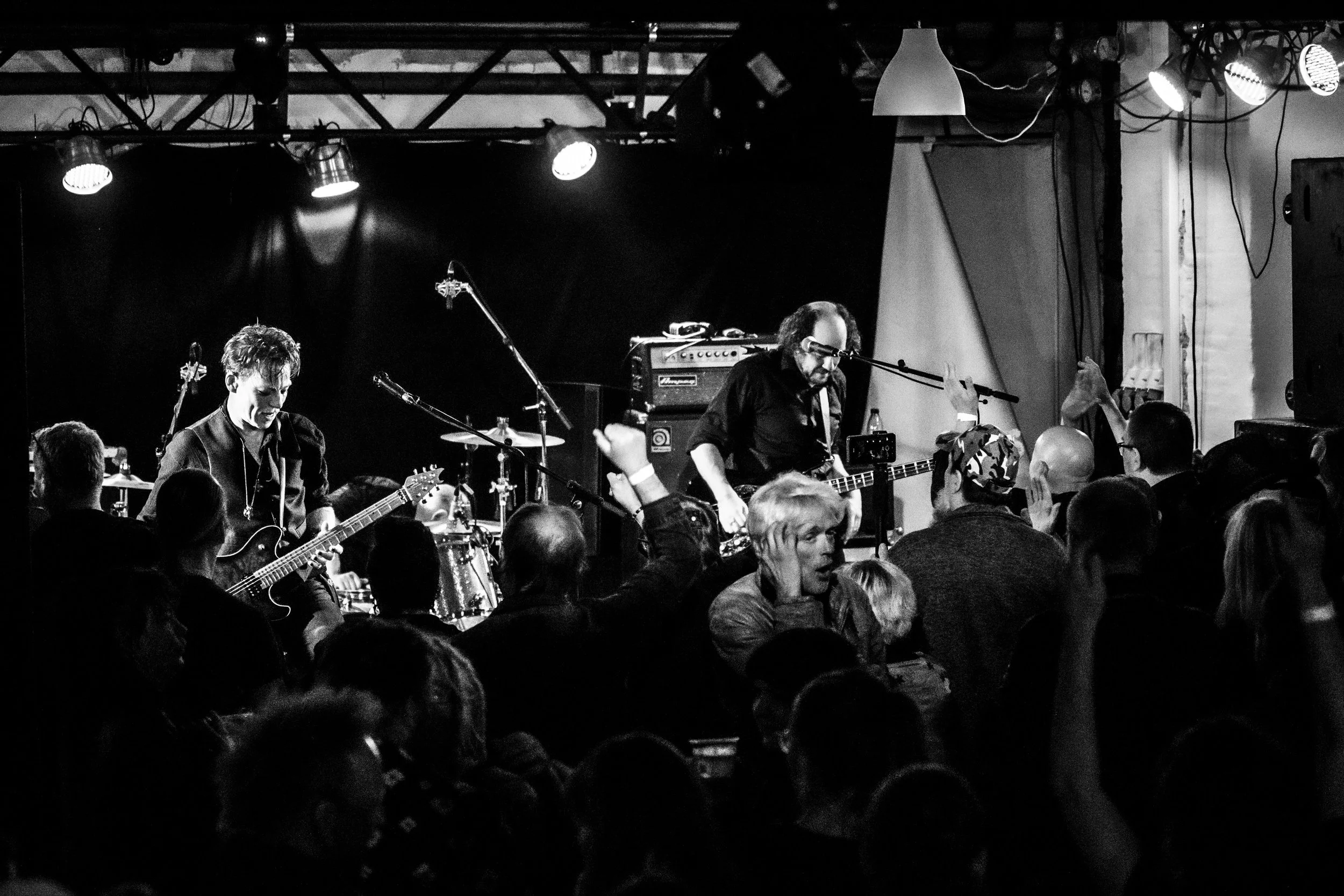 Black and white photo of a live concert with a band playing on stage. The audience is visible, some with hands raised, and the band members are singing and playing guitars.