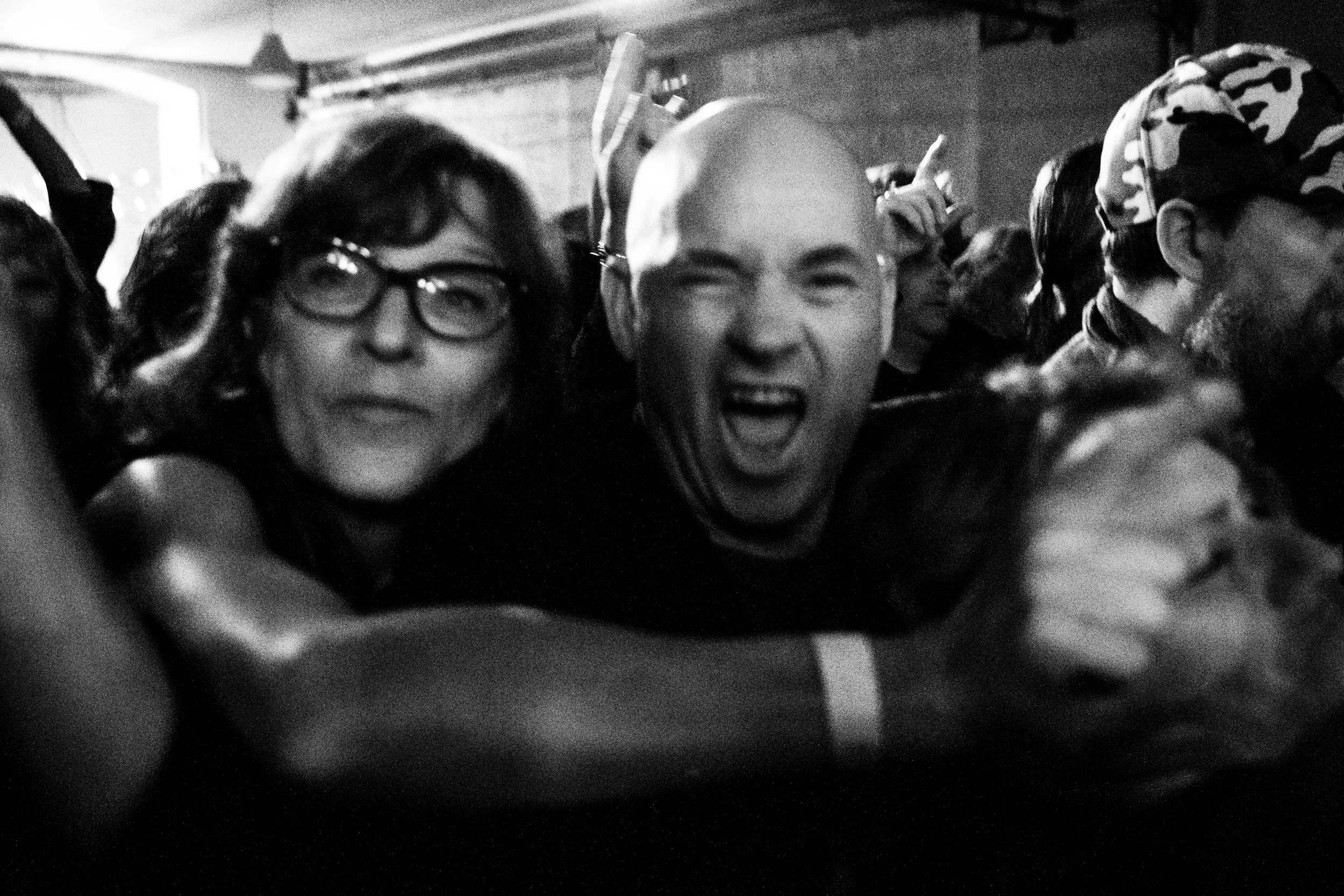 A group of people at a lively event, with focus on a bald man smiling and a woman with glasses, all showing excitement.