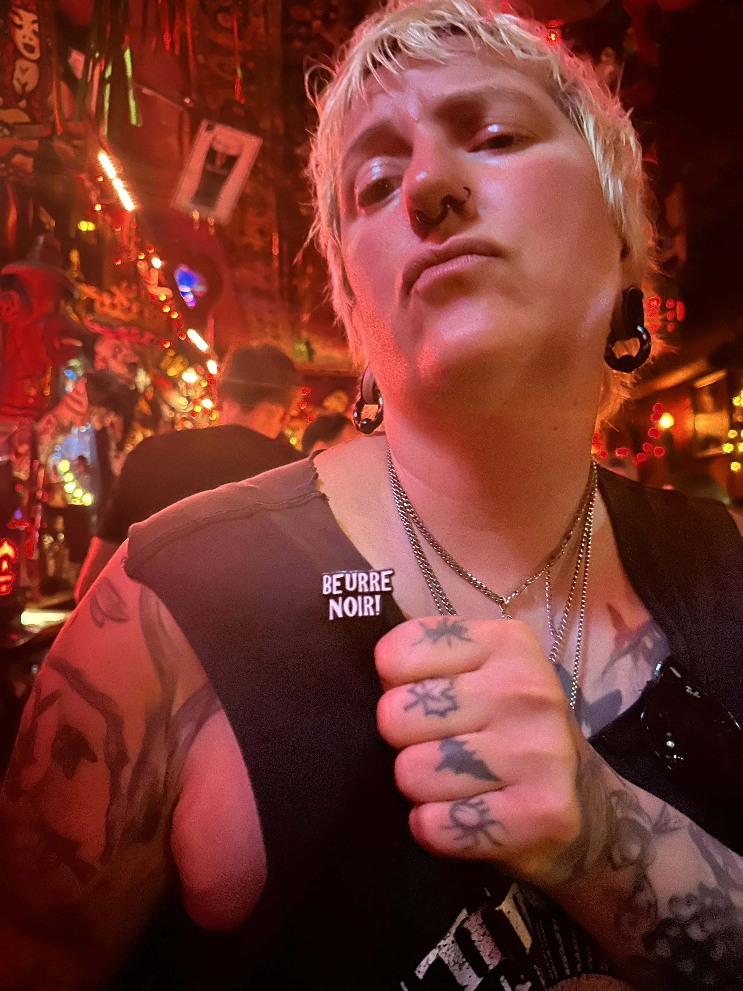 Close-up of a person with short blond hair, tattoos, piercings, and wearing black earrings and layered necklaces, in a colorful, neon-lit bar or club.