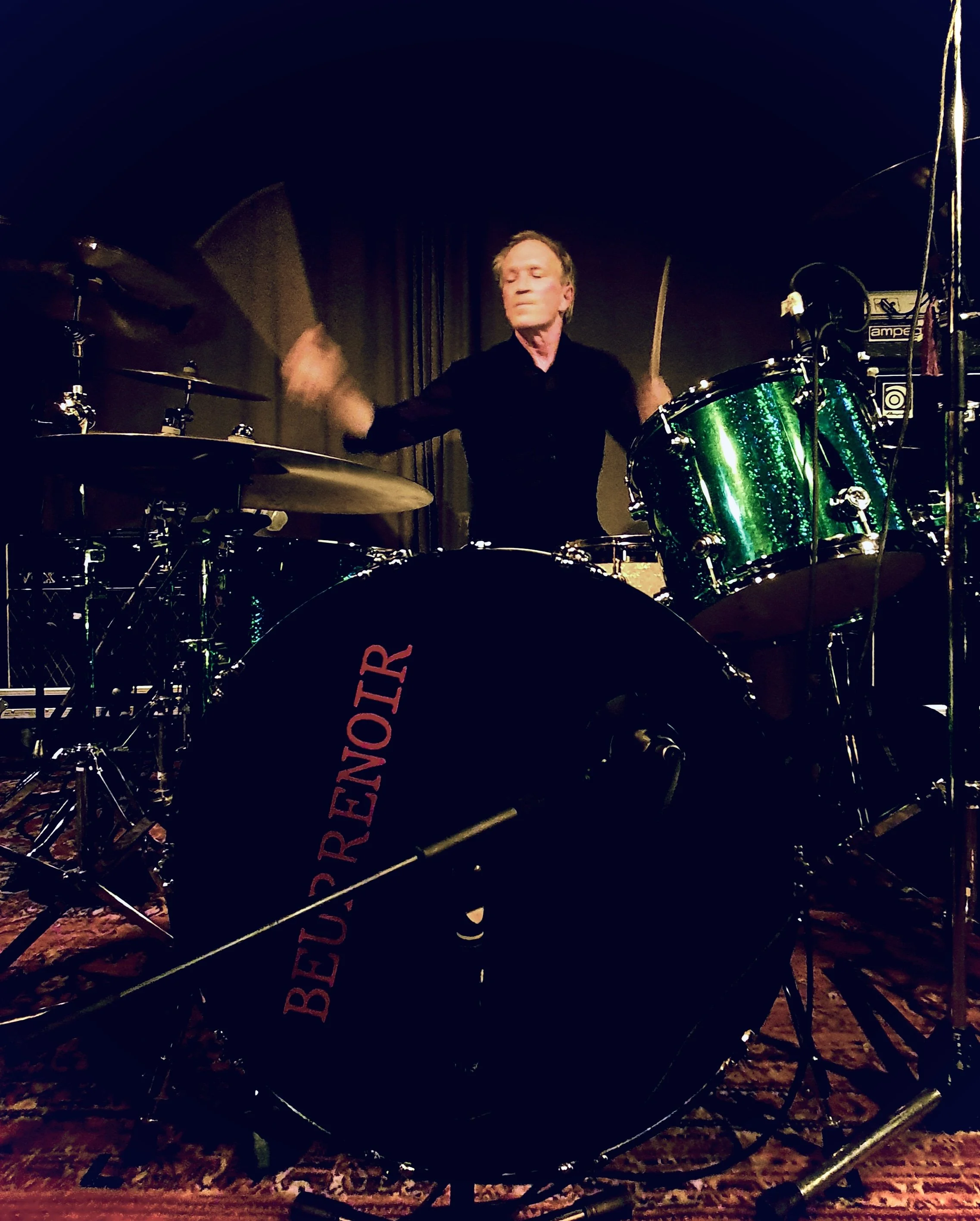 A man playing the drums on a stage, with a black drum set and green sparkle drums, in a dimly lit environment.