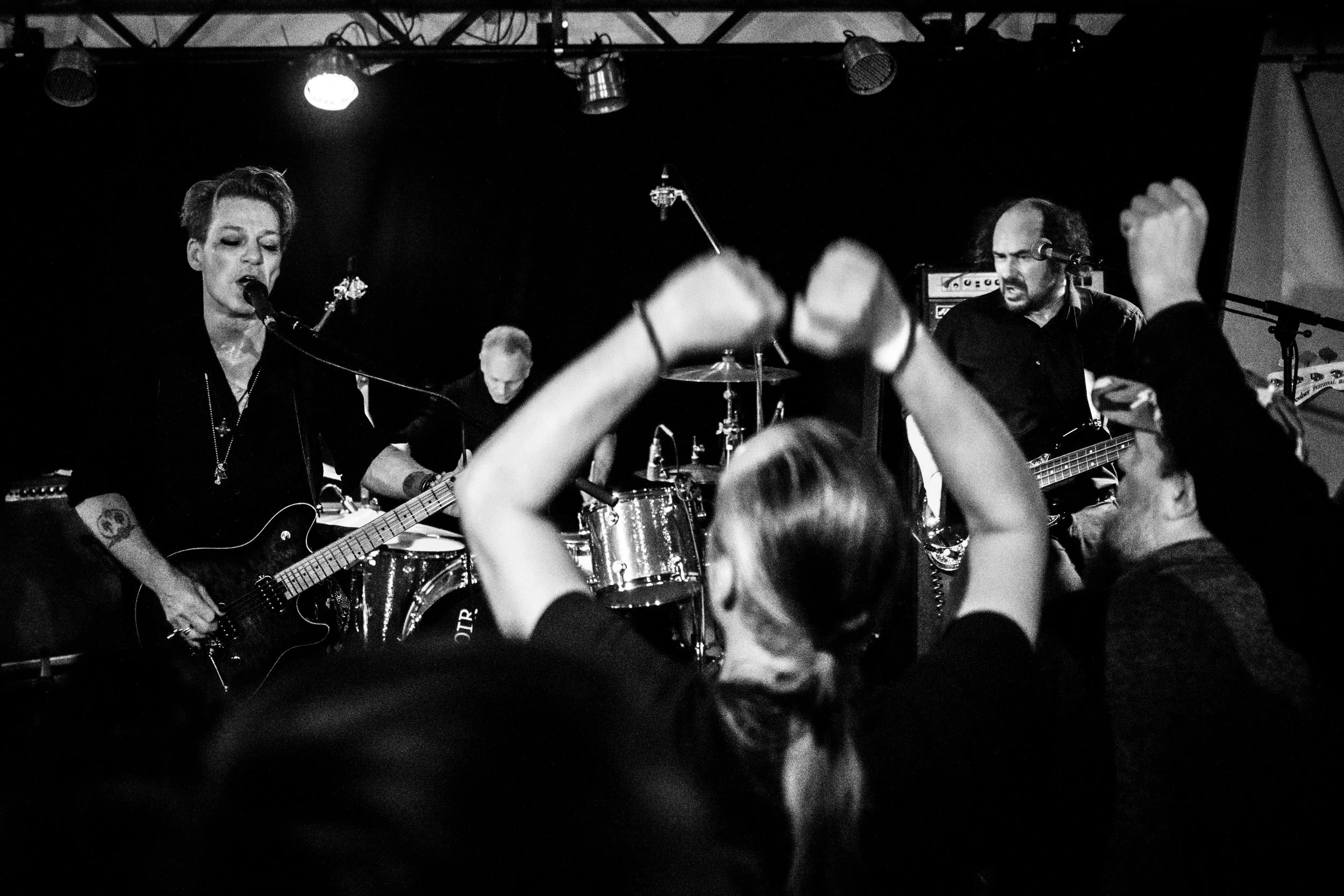 A black-and-white photo of a band performing live on stage with audience members raising their arms in the foreground.