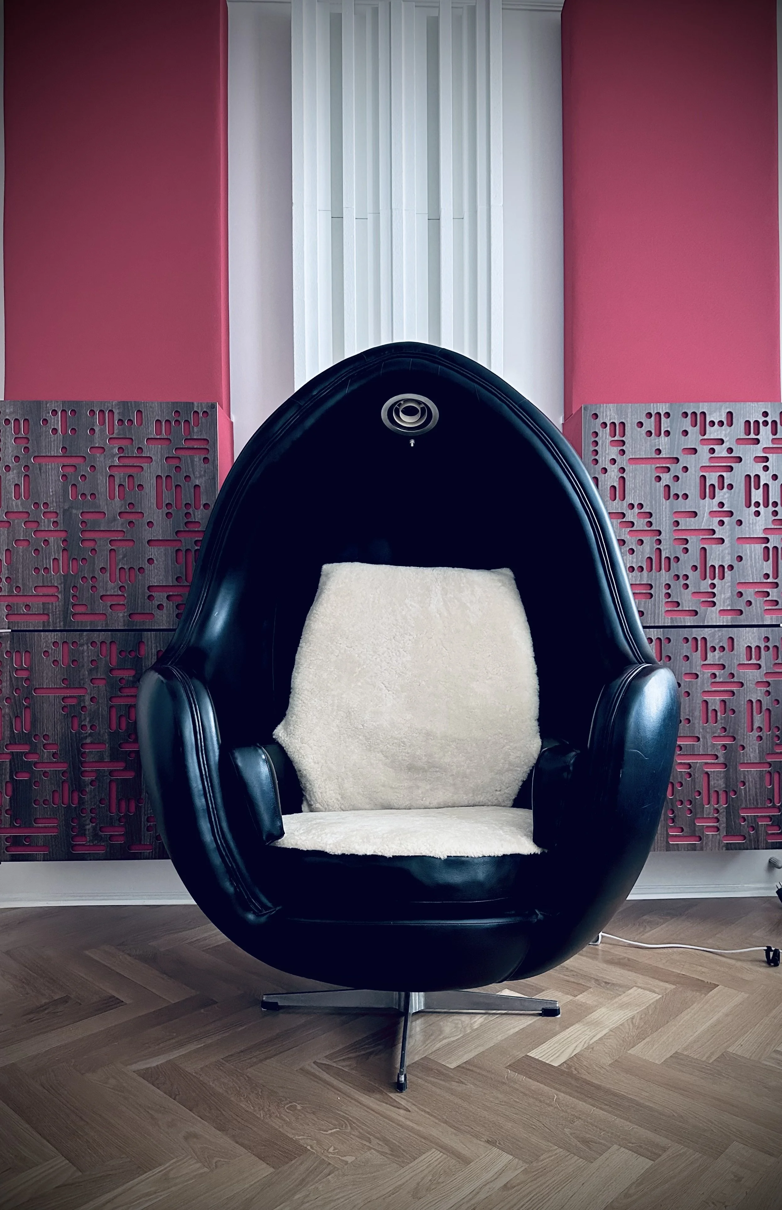 Black egg-shaped chair with a cream-colored cushion inside, set against a pink and white panel wall and wooden floor.