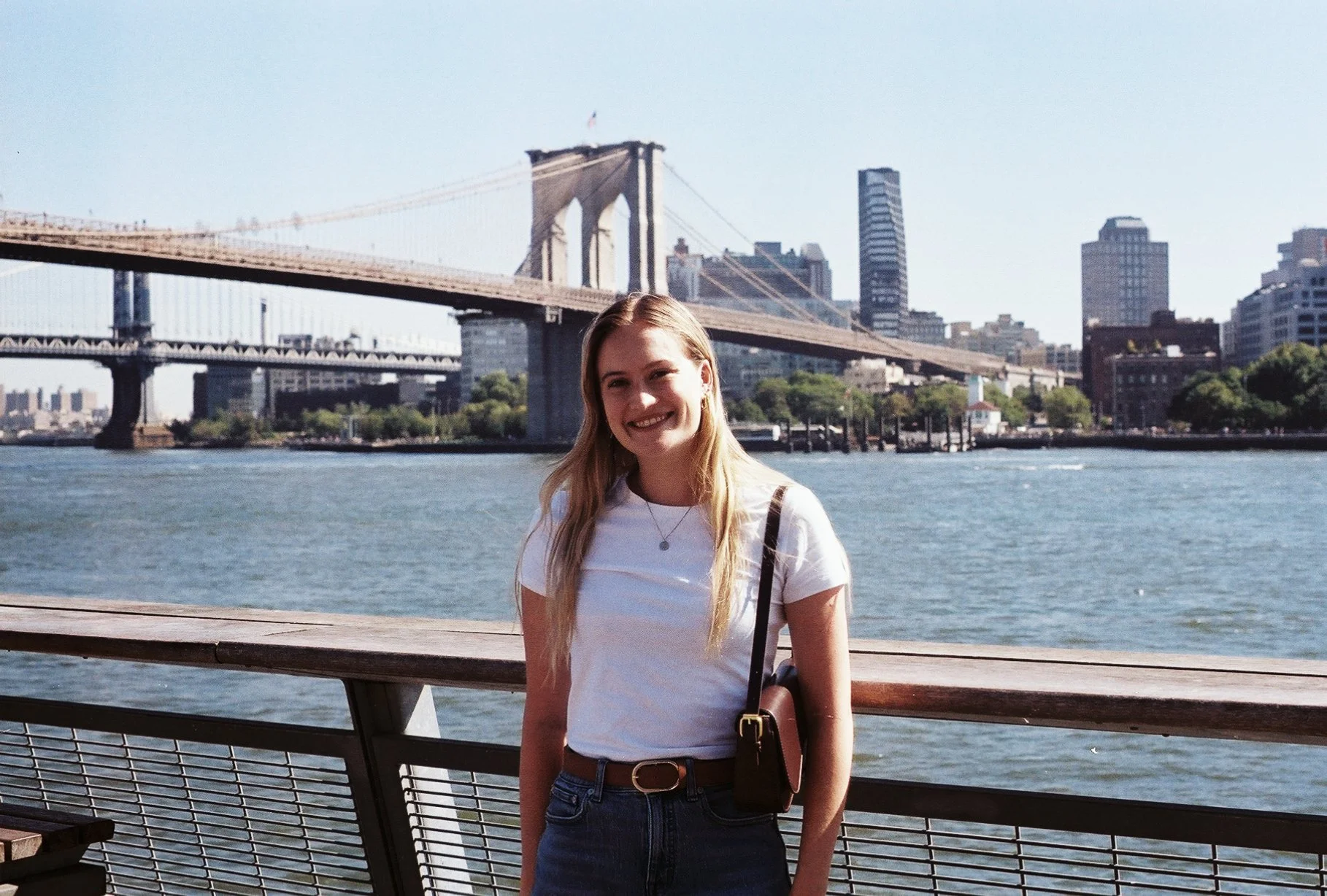 Young woman with long blonde hair, wearing a white t-shirt, jeans, and carrying a purse, standing on a waterfront with the Brooklyn Bridge and Manhattan skyline in the background.
