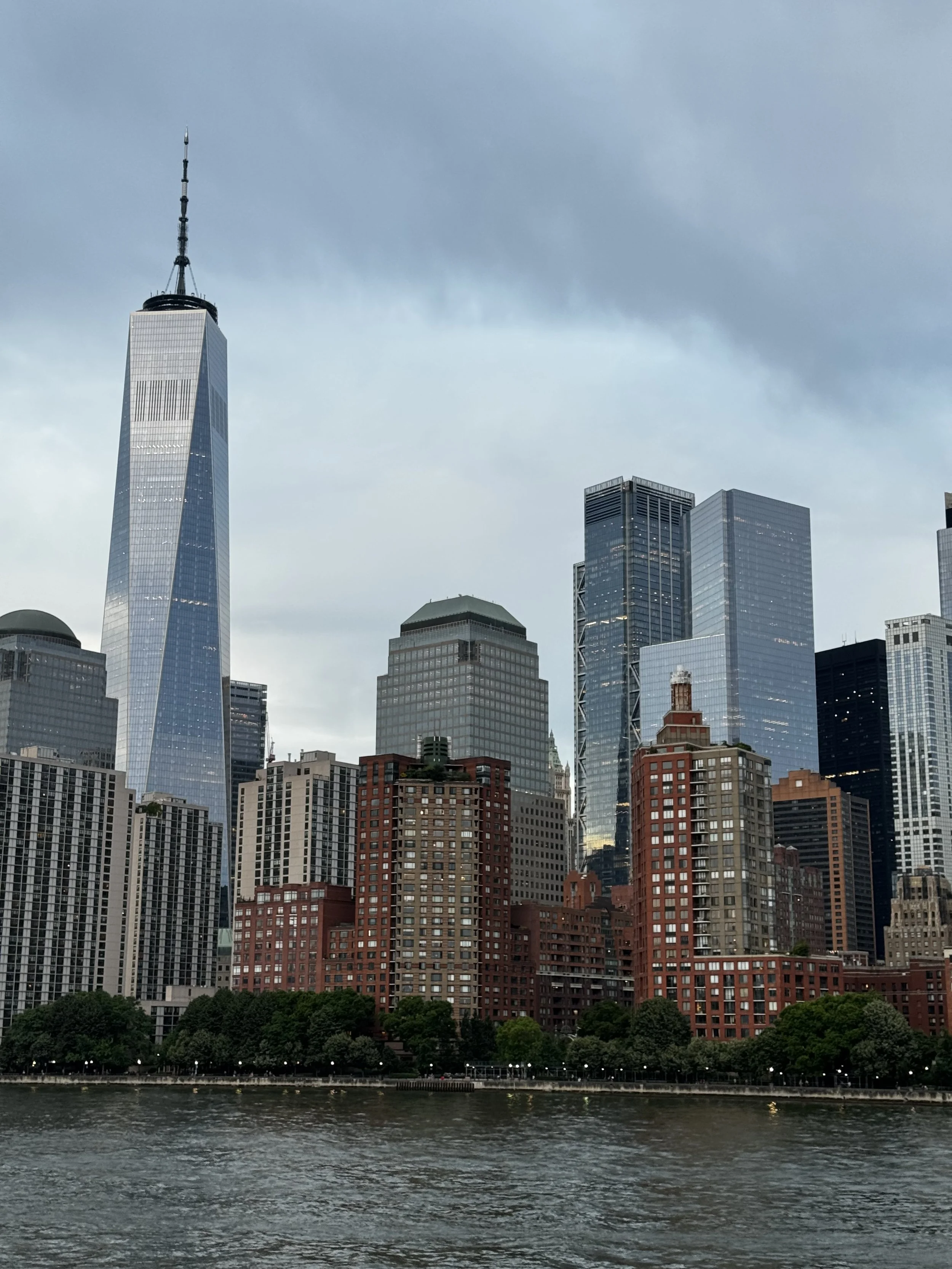 A city skyline with tall skyscrapers, including One World Trade Center, viewed from across a body of water with a tree-lined shoreline in the foreground.