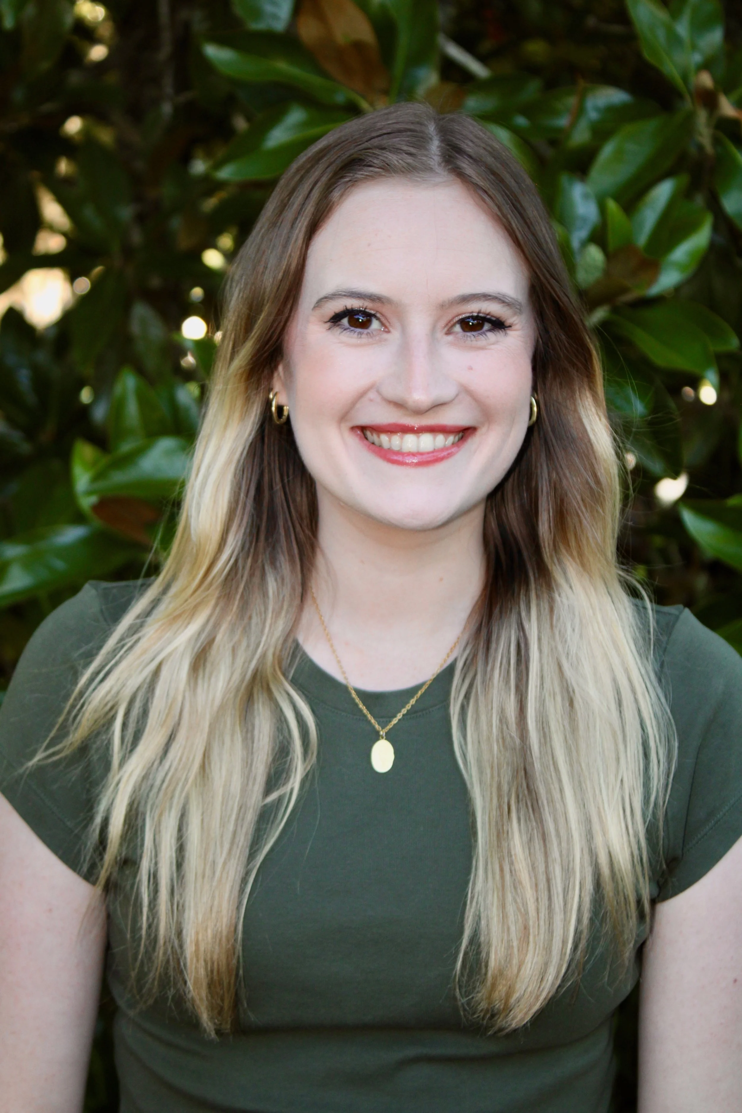Portrait of a young woman with long, wavy hair smiling, wearing a green top, gold hoop earrings, and necklaces, standing in front of a leafy background.