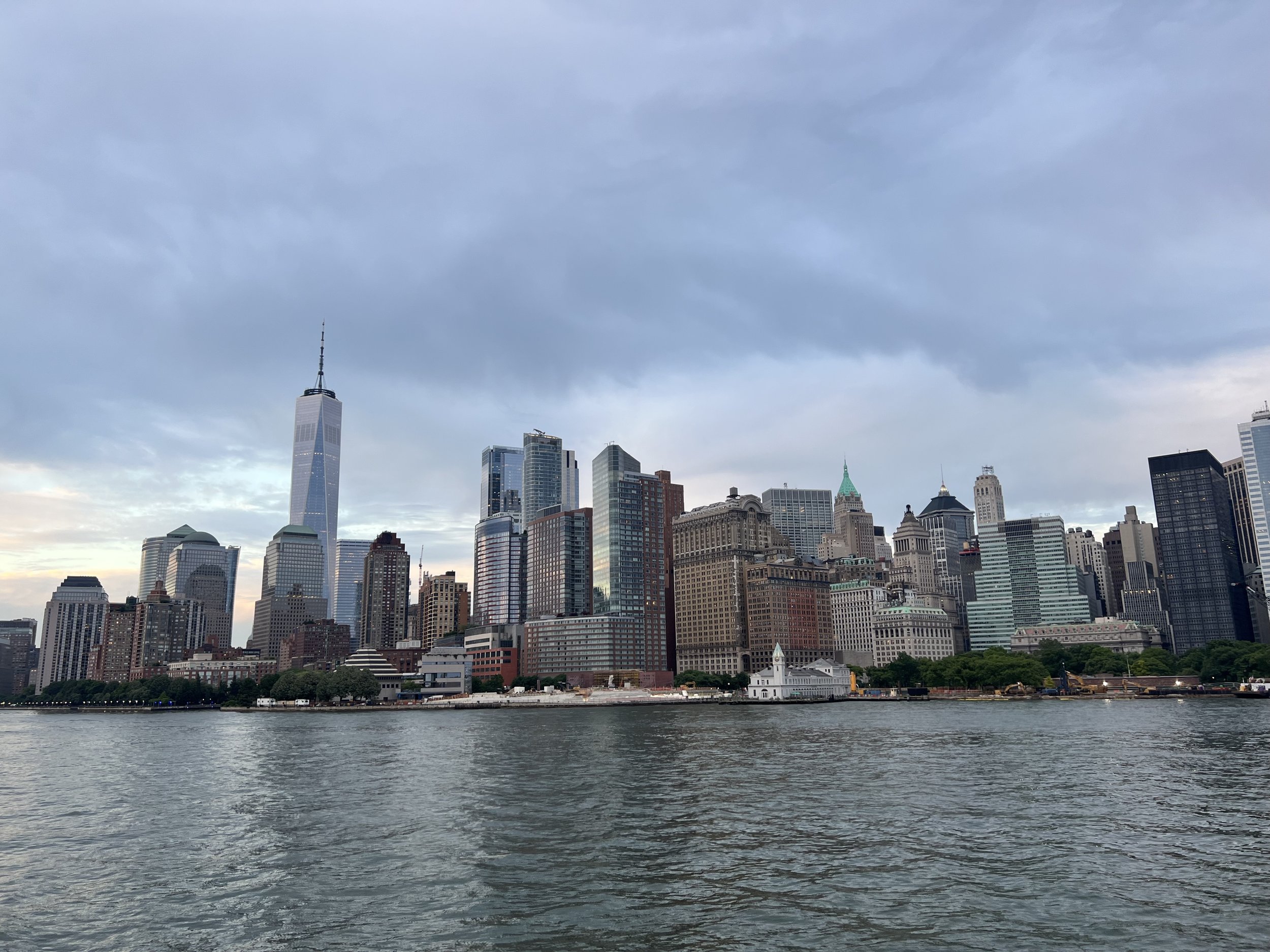 A city skyline viewed from across a body of water, featuring tall skyscrapers including One World Trade Center, under a cloudy sky.