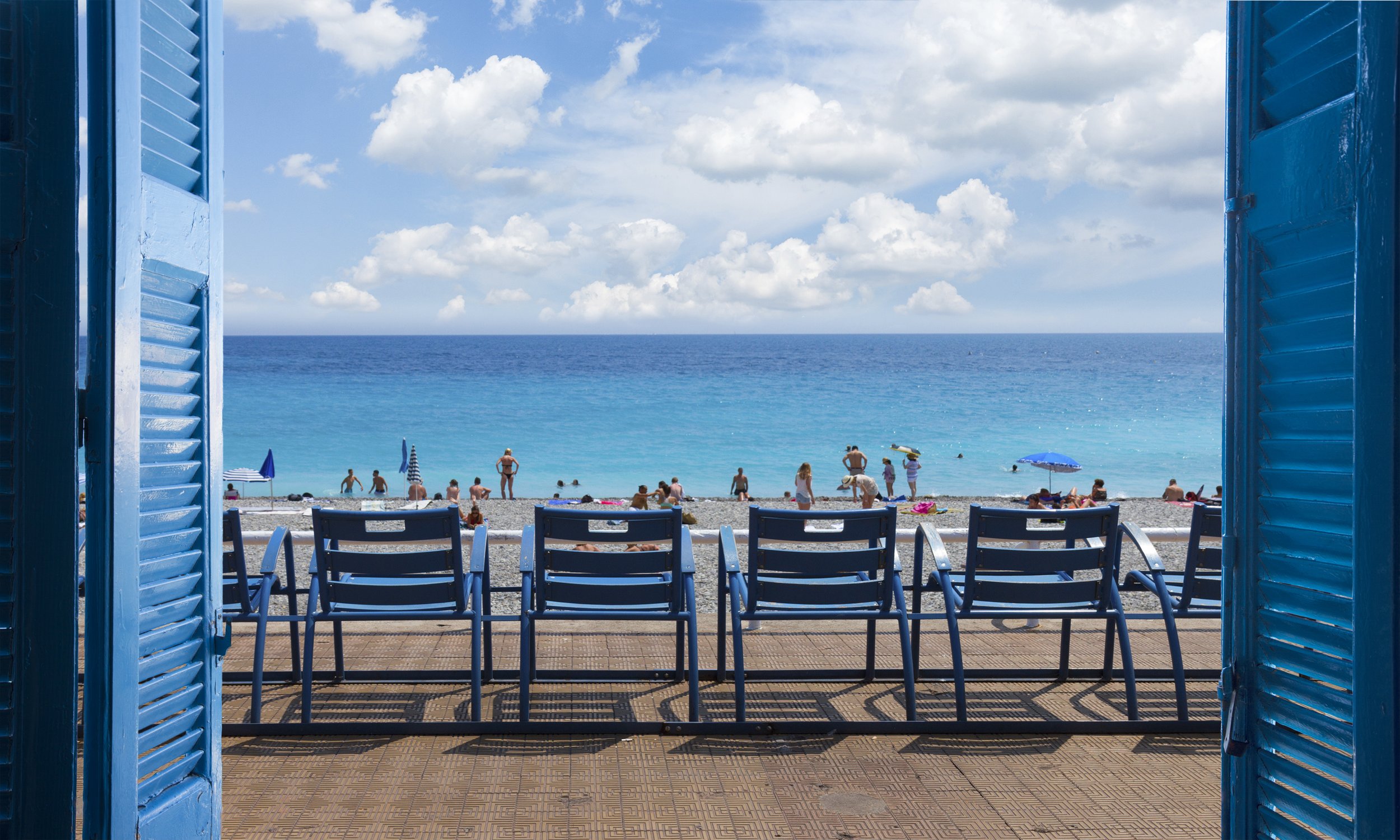 View of a beach in Nice, France, seen through blue shuttered window, with empty blue chairs facing people sunbathing, swimming, and relaxing under umbrellas, and the ocean and sky in the background.