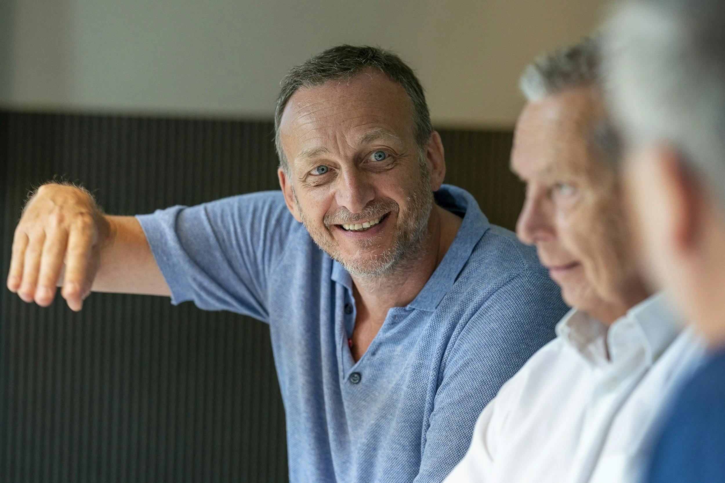 A man with blue eyes, short gray hair, and a beard wearing a blue shirt, smiling and pointing at a woman with gray hair and a white blouse.