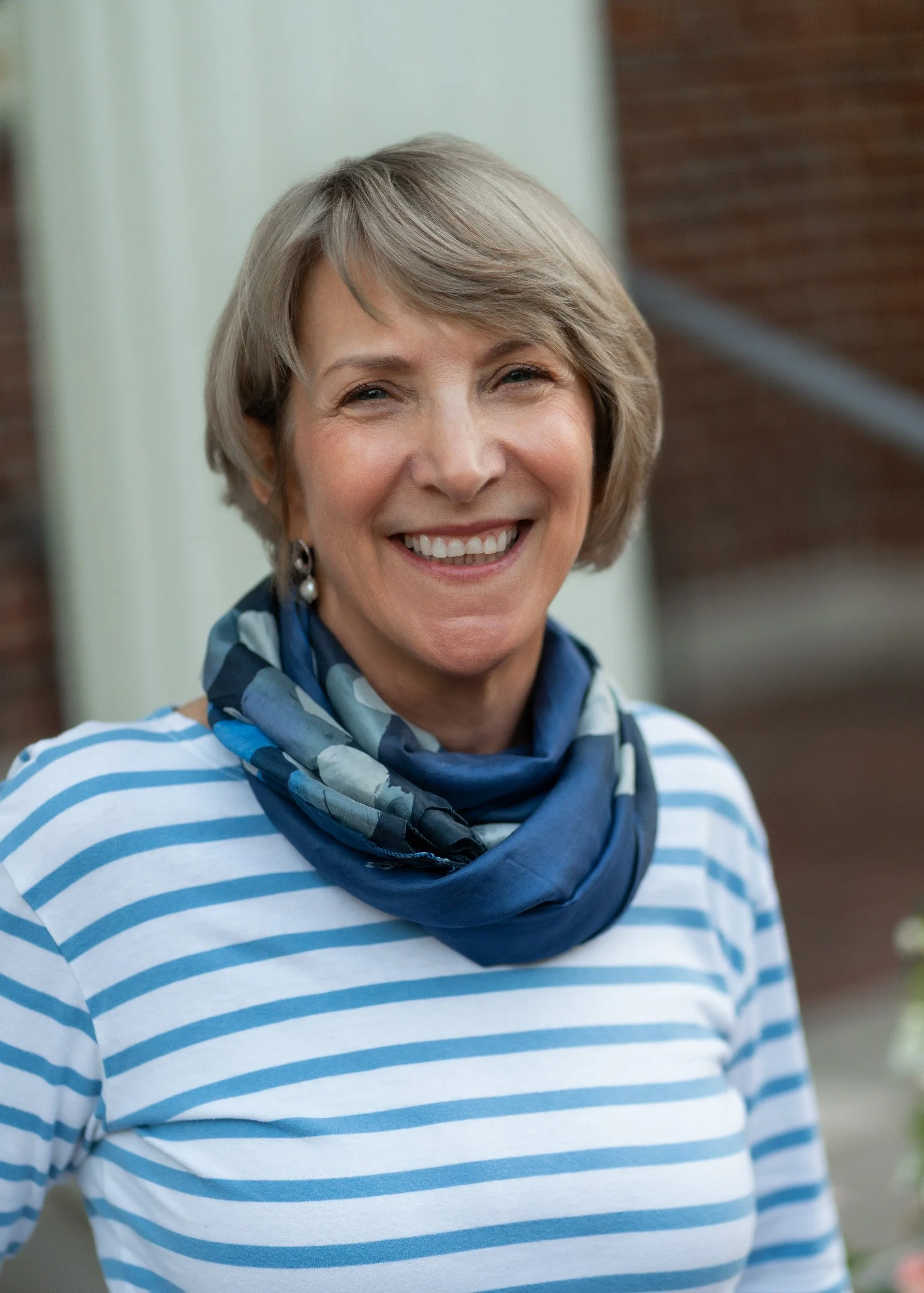 Close-up of a smiling older woman with short gray hair, wearing a blue and white striped shirt and a blue patterned scarf.
