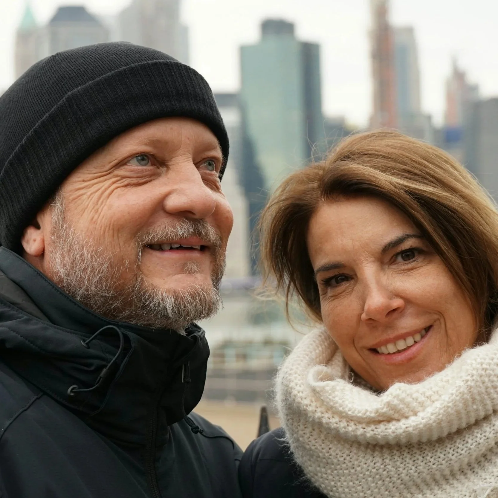 A close-up of a man in a black beanie and a woman with a knitted cream-colored scarf, both smiling and standing outdoors with a city skyline in the background.