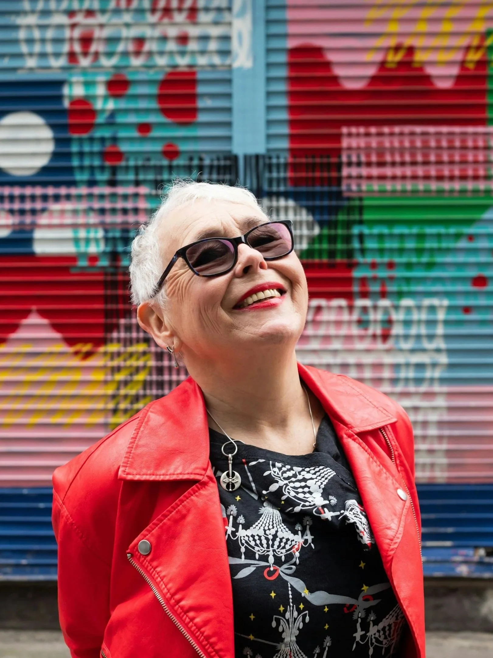 An older woman with short white hair, sunglasses, red lipstick, and a red jacket smiling in front of colorful graffiti on a metal wall.
