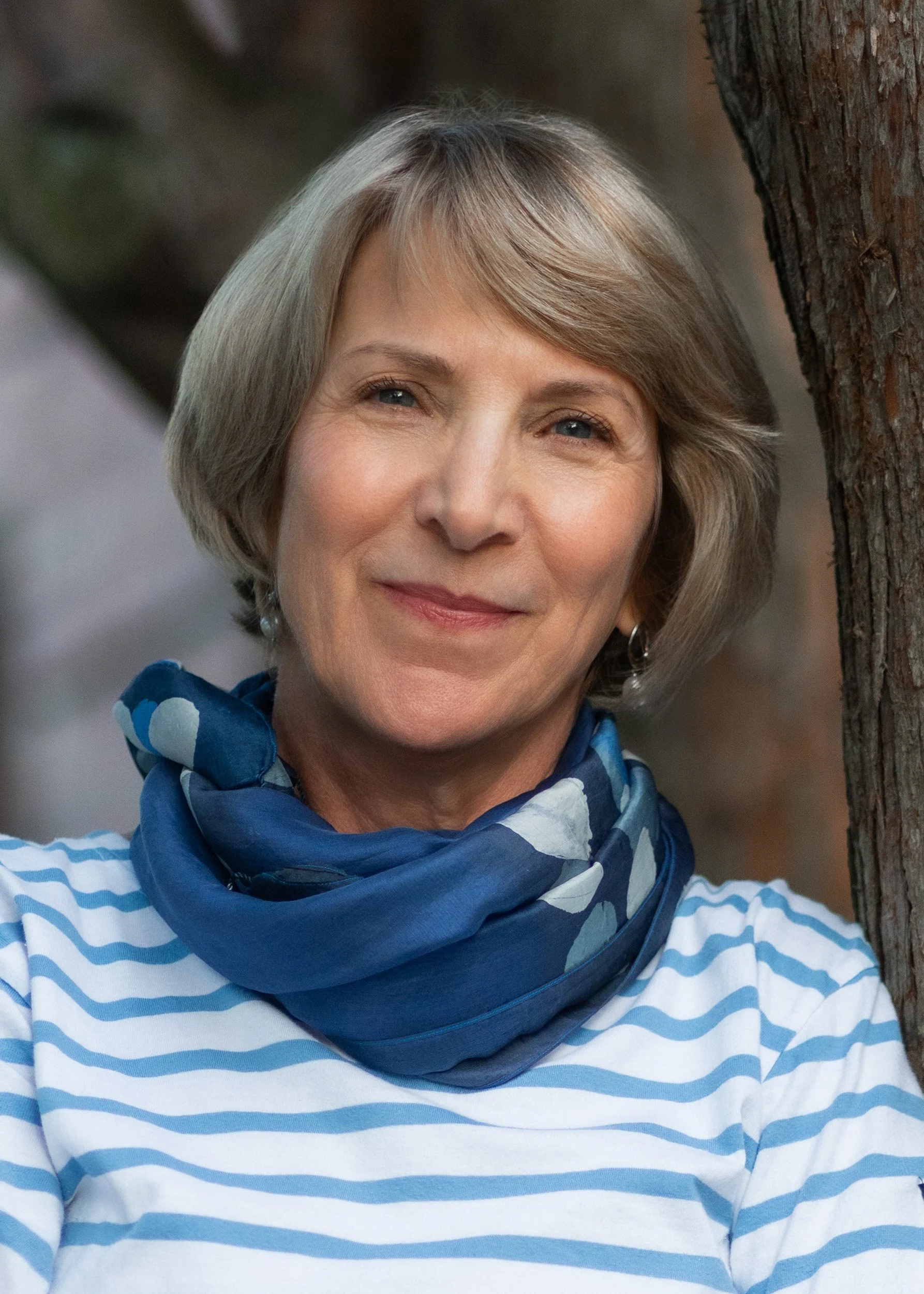 A smiling woman with short brown hair and blue eyes, wearing a navy blue scarf and a white and blue striped shirt, standing outdoors near a tree.