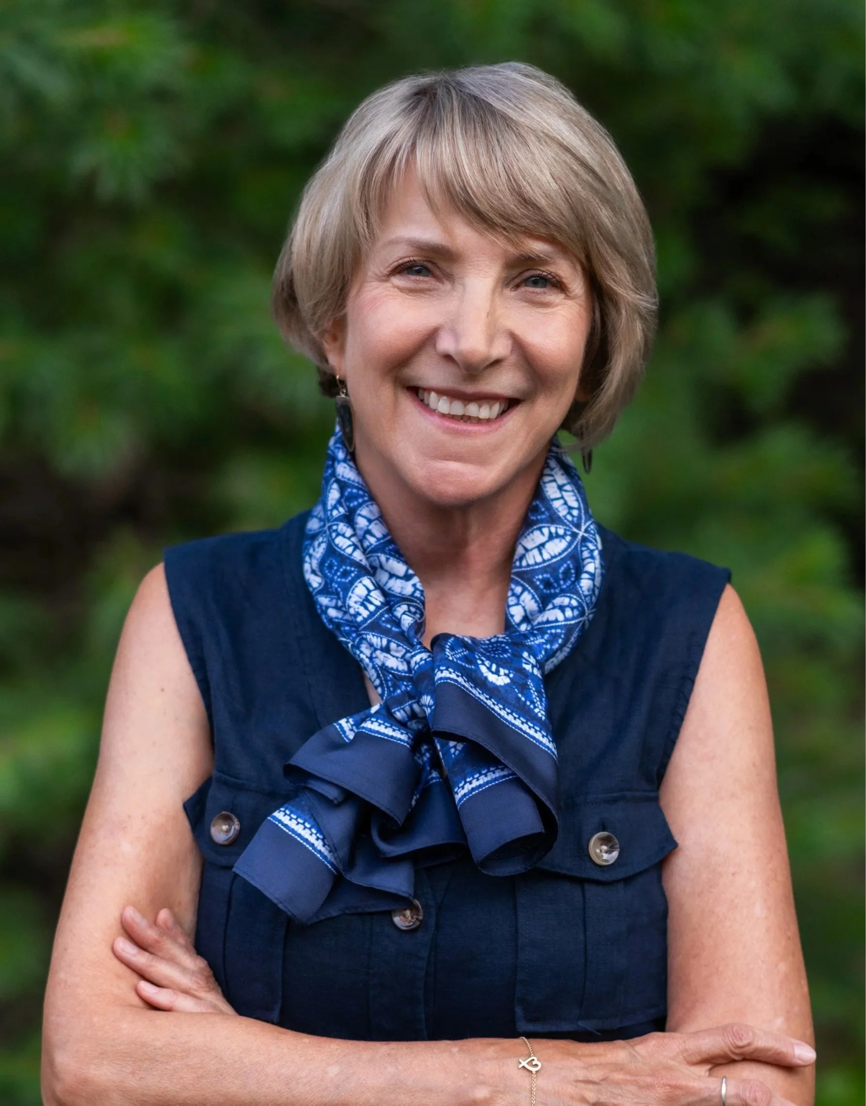 A middle-aged woman with short blonde hair smiling outdoors, wearing a navy vest with gold buttons and a blue patterned scarf. She has a gold bracelet with an infinity symbol.