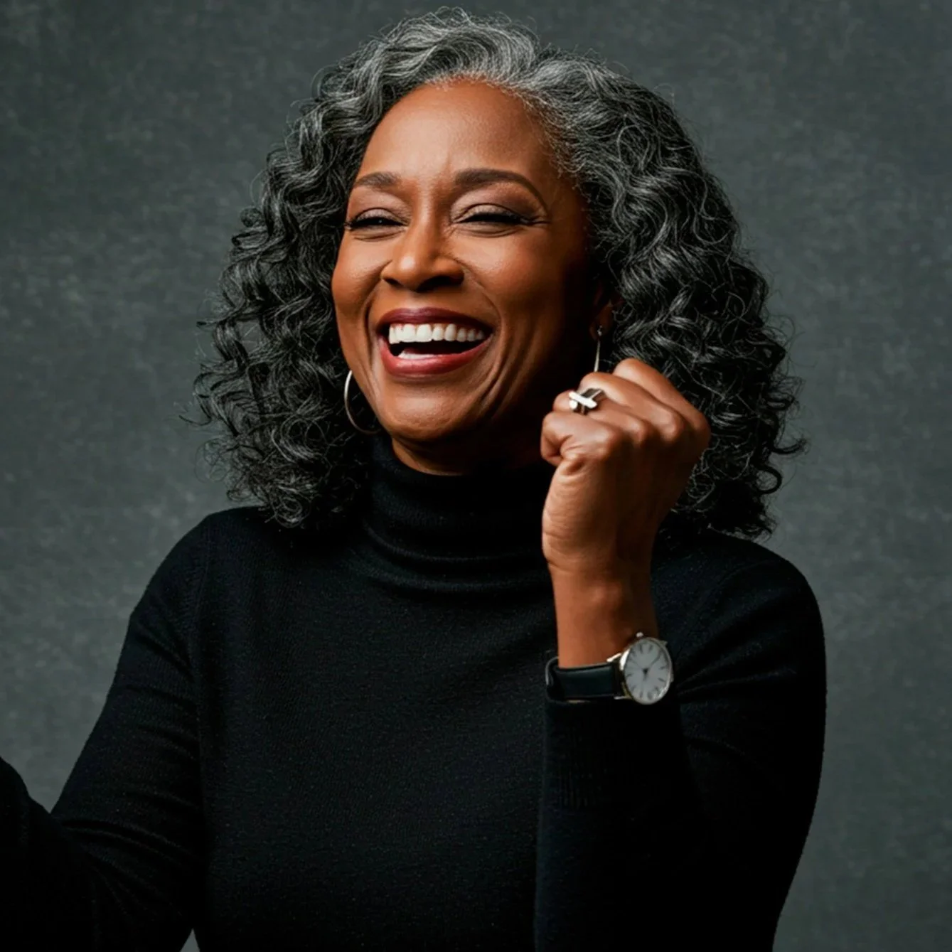 Smiling mature woman with gray curly hair, wearing a black turtleneck, silver earrings, and a watch, against a dark background.