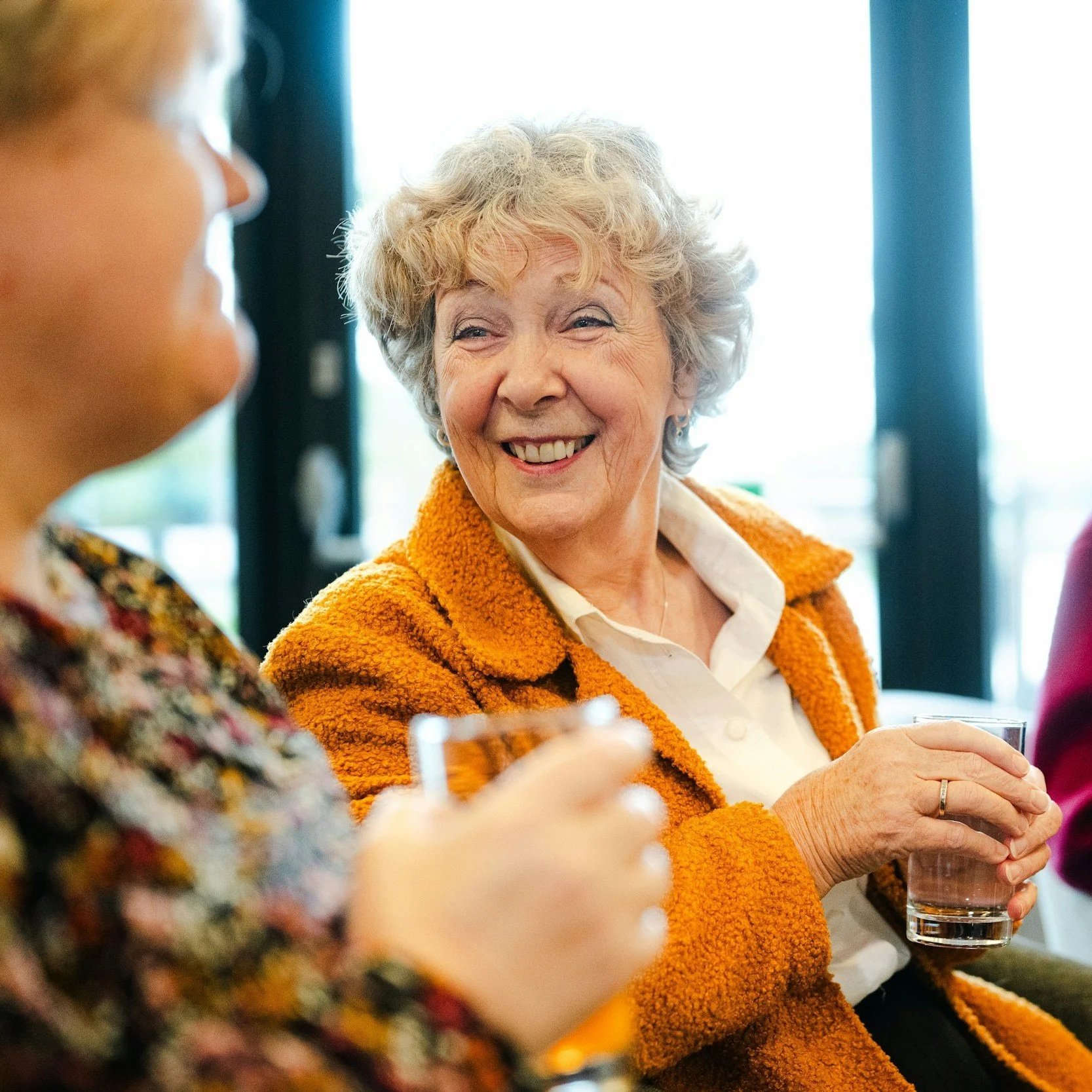 A senior woman with curly gray hair smiling and holding a glass of water, engaged in conversation with a person wearing a colorful patterned top.