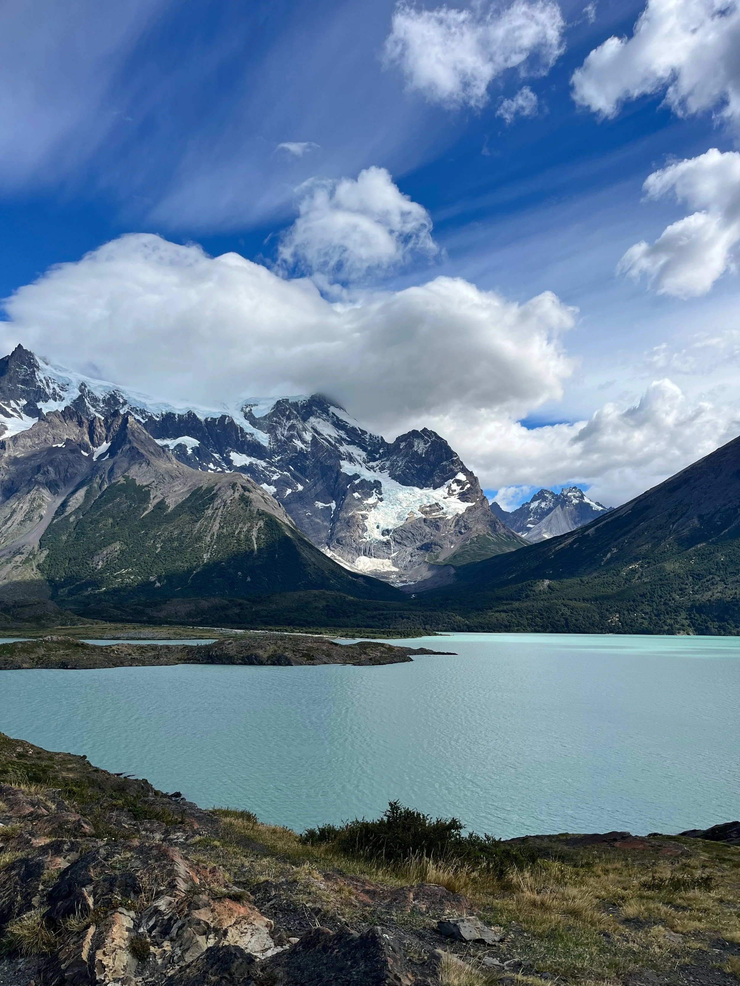A mountain landscape in Chile with snow-capped peaks, a glacier, green forested slopes, and a turquoise lake in the foreground under a partly cloudy sky.