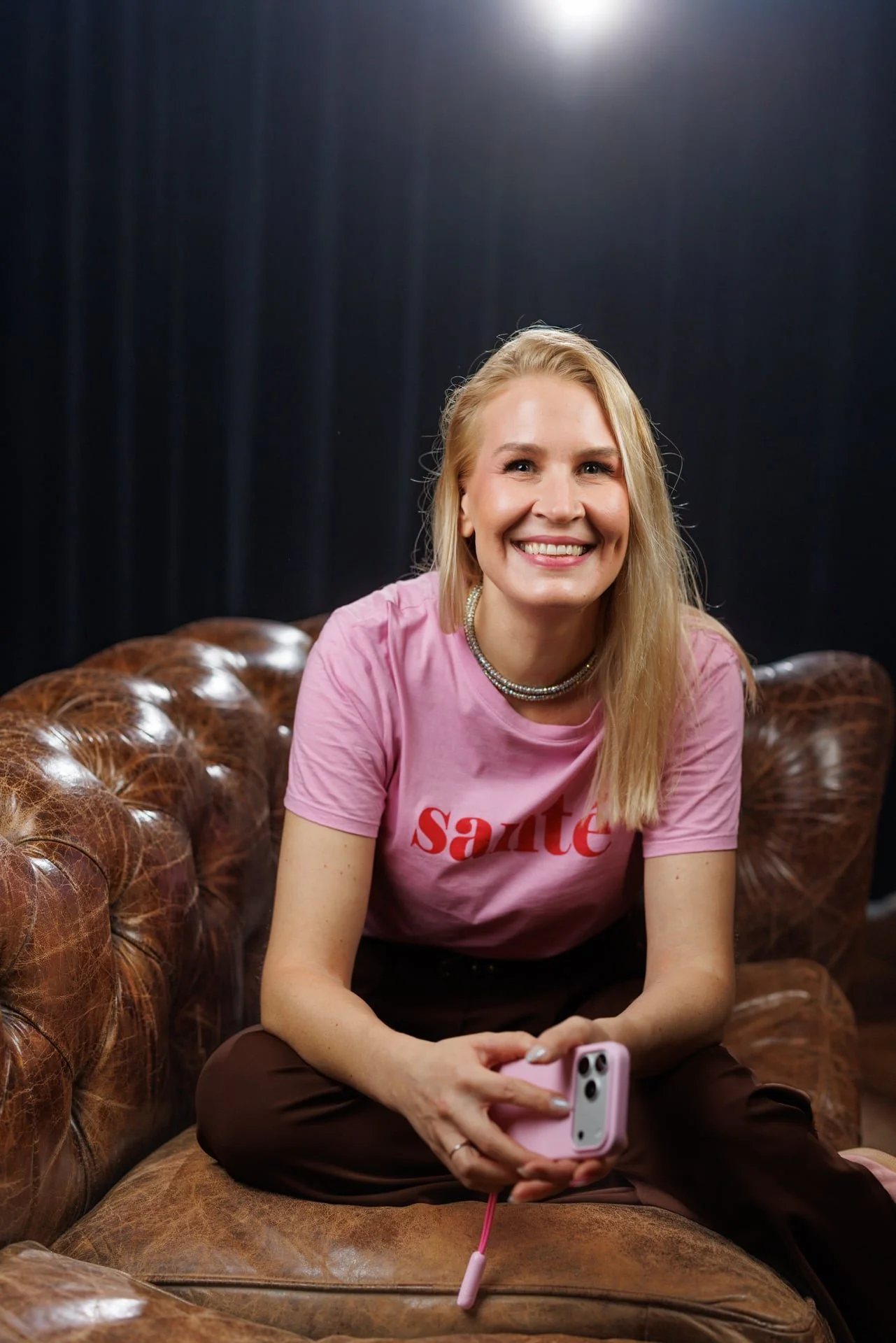 Meri Wiik har blondt hår, er iført en rosa t-skjorte og brune bukser. Hun sitter i en skinnsofa, smiler til kameraet og holder en rosa smarttelefon. Meri Wiik er ekspert på merkevare, LinkedIn og ansatte i sosiale medier.