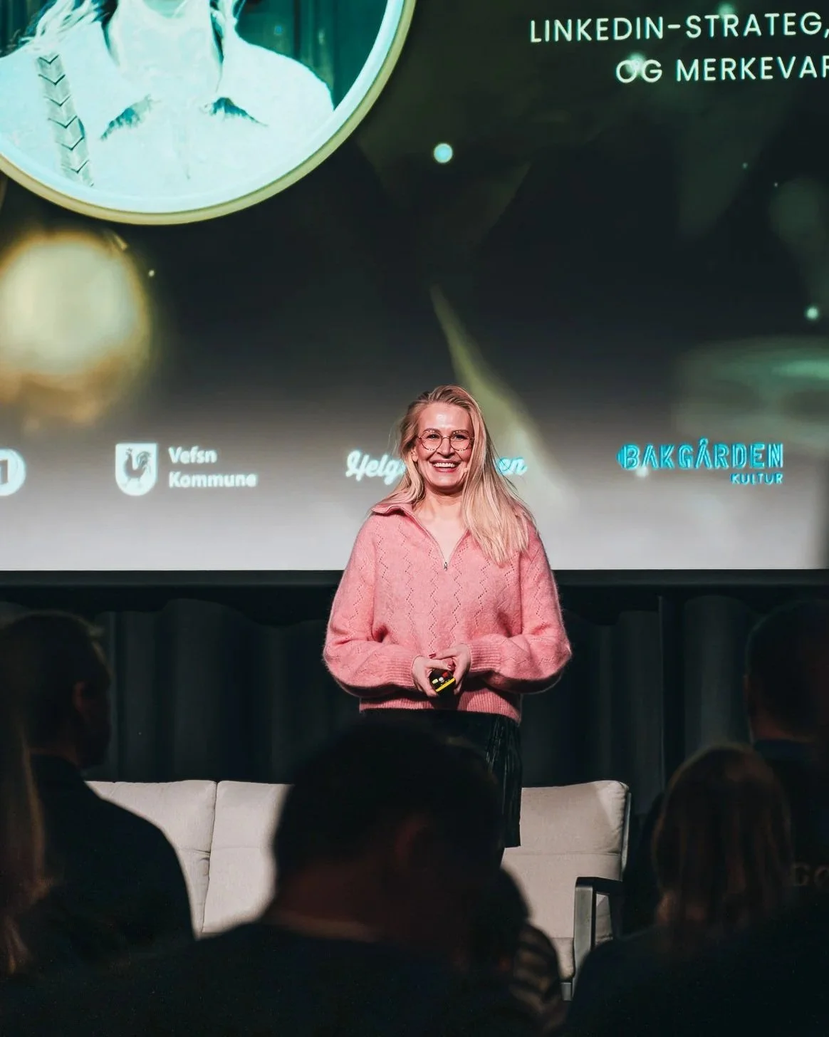 Meri Wiik med langt blondt hår, briller og en rosa genser står på scenen med en mikrofon i hånden, smilende, foran en skjerm som viser en presentasjon om LinkedIn og merkevarebygging for bedrift.