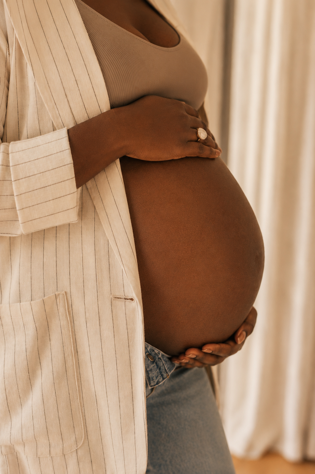 Close-up of a pregnant woman cradling her belly with her hands, wearing a beige tank top and a beige pinstriped blazer.