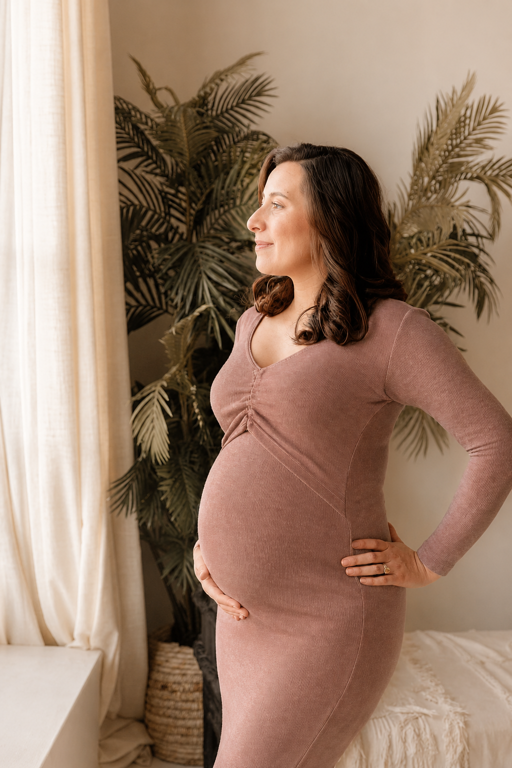 Pregnant woman standing near window, gazing outside, hands on hips, in a cozy room with beige curtains and green plants in the background.