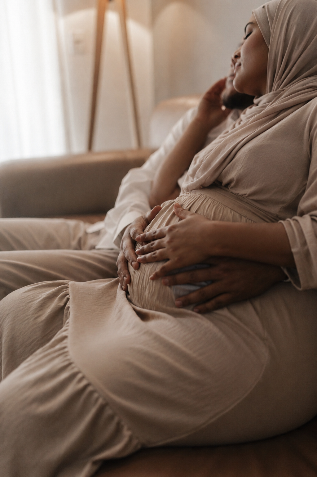 A woman in a beige dress and headscarf sitting on a couch, holding her pregnant belly with one hand, with another person behind her also touching her belly.