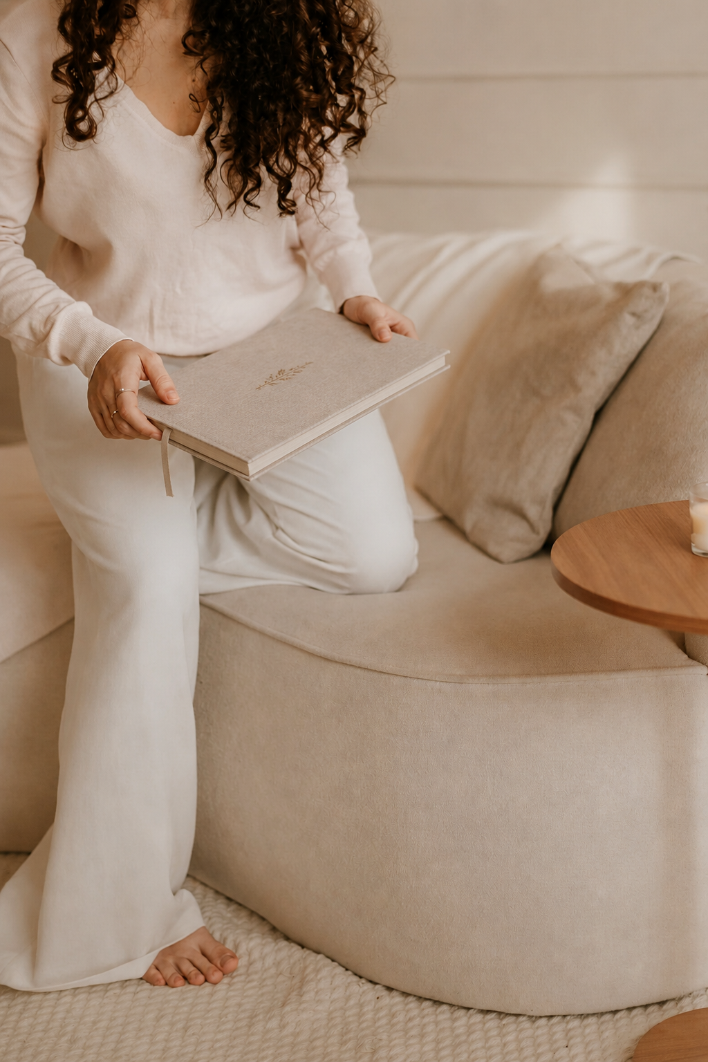 A woman in white cozy outfit sitting on a beige sofa, holding a grey book with a small gold design, next to a wooden side table with a candle, and beige pillows on the sofa.