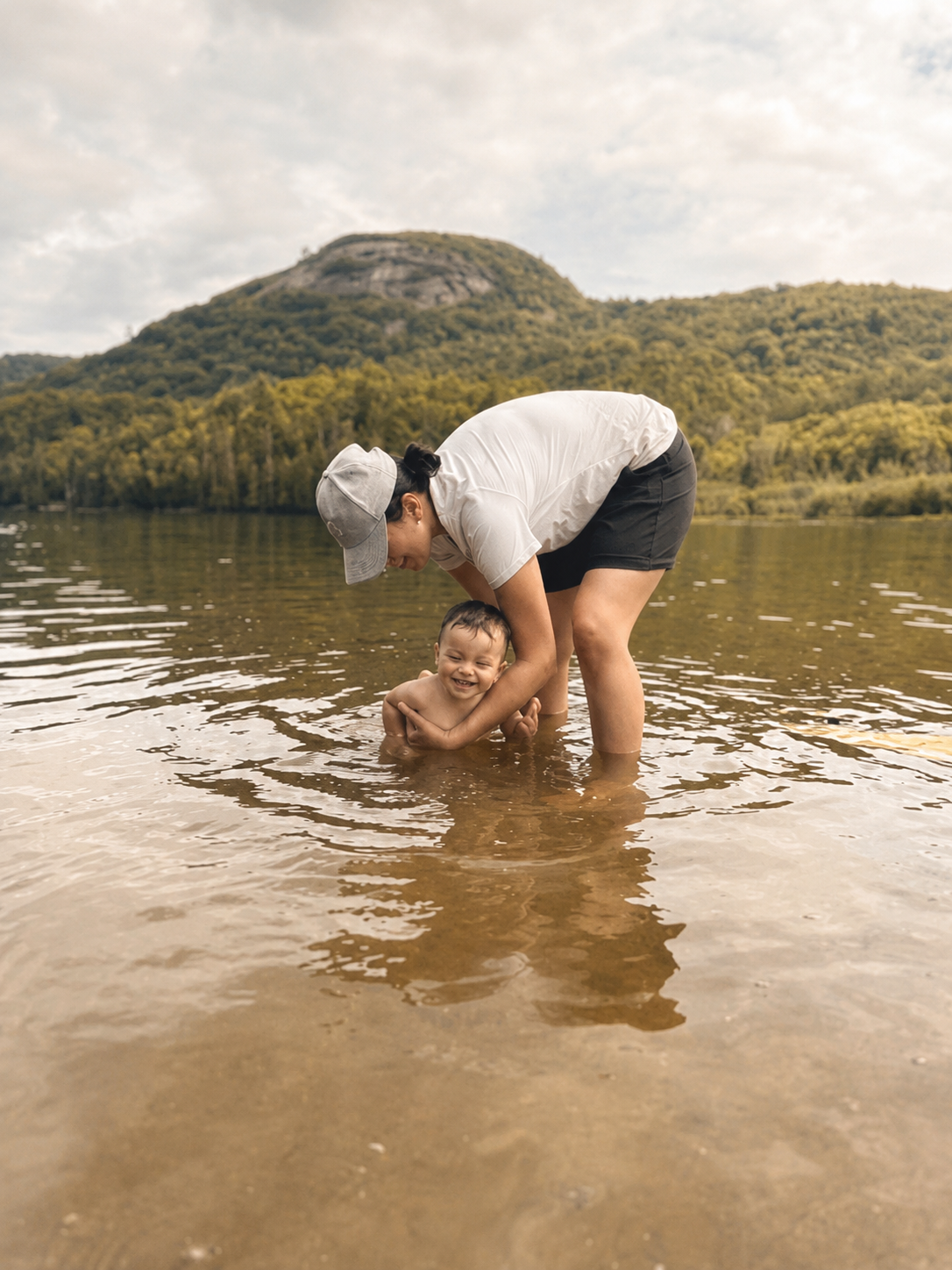 A woman and a young boy playing and splashing in a shallow river, surrounded by nature and mountains.