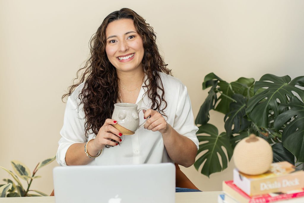 A woman with long curly brown hair smiling, holding a ceramic mug, sitting at a desk with a laptop, surrounded by indoor plants and books.