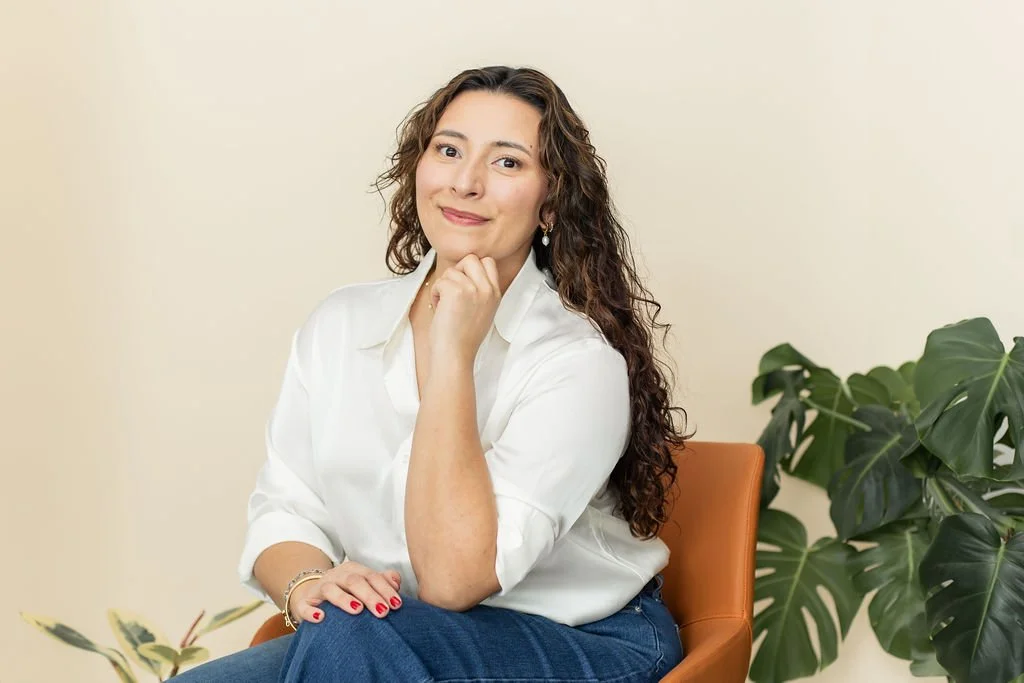 A woman with curly hair sitting on a brown chair, wearing a white blouse and blue jeans, smiling with her right hand resting on her chin, next to a green leafy plant.