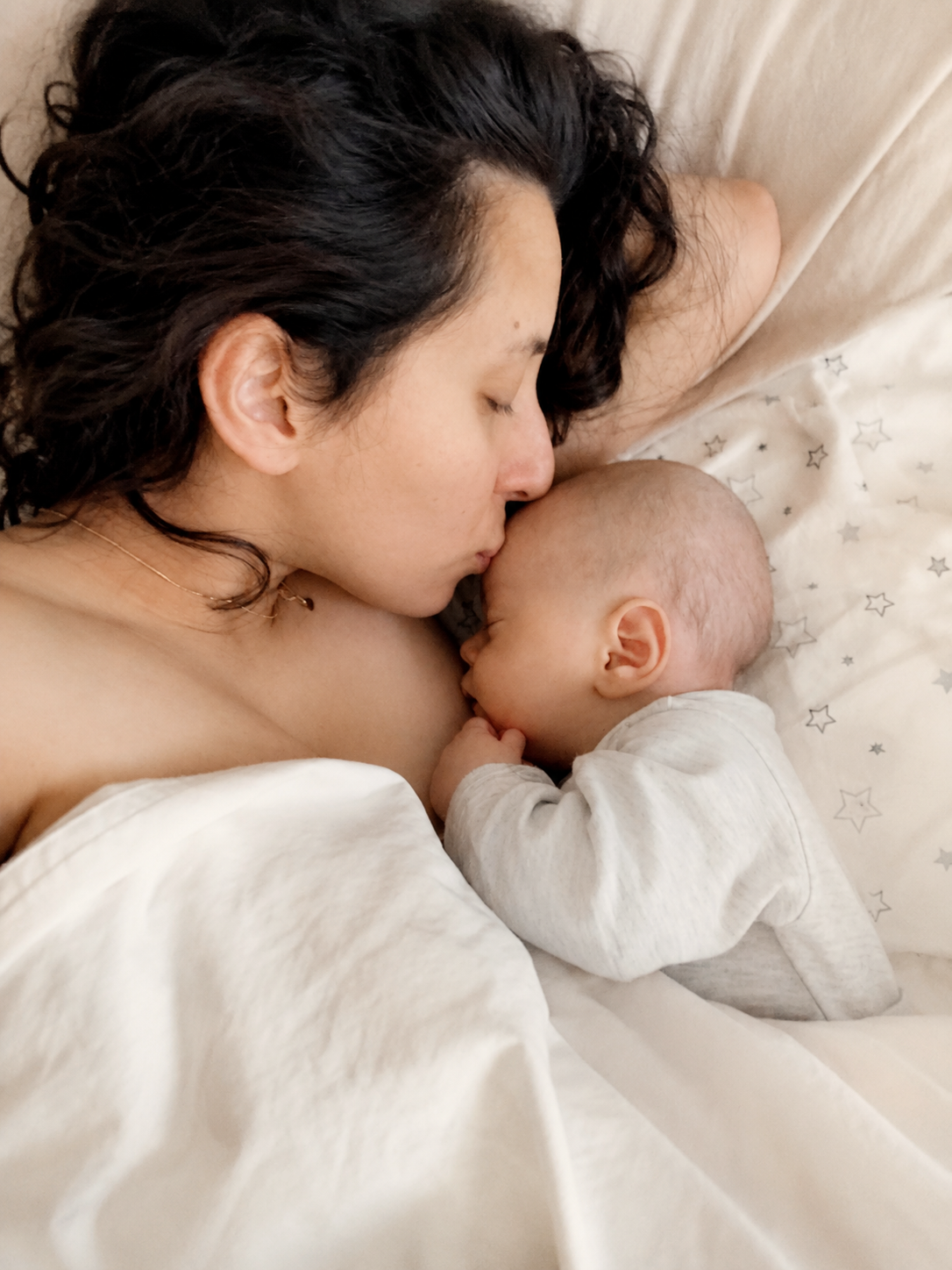 A woman with dark, wavy hair sleeping on a bed, kissing a small baby on the forehead. The baby, wearing a light gray outfit, is also sleeping with their hand near their face. They are on a bed with white sheets that have a star pattern.
