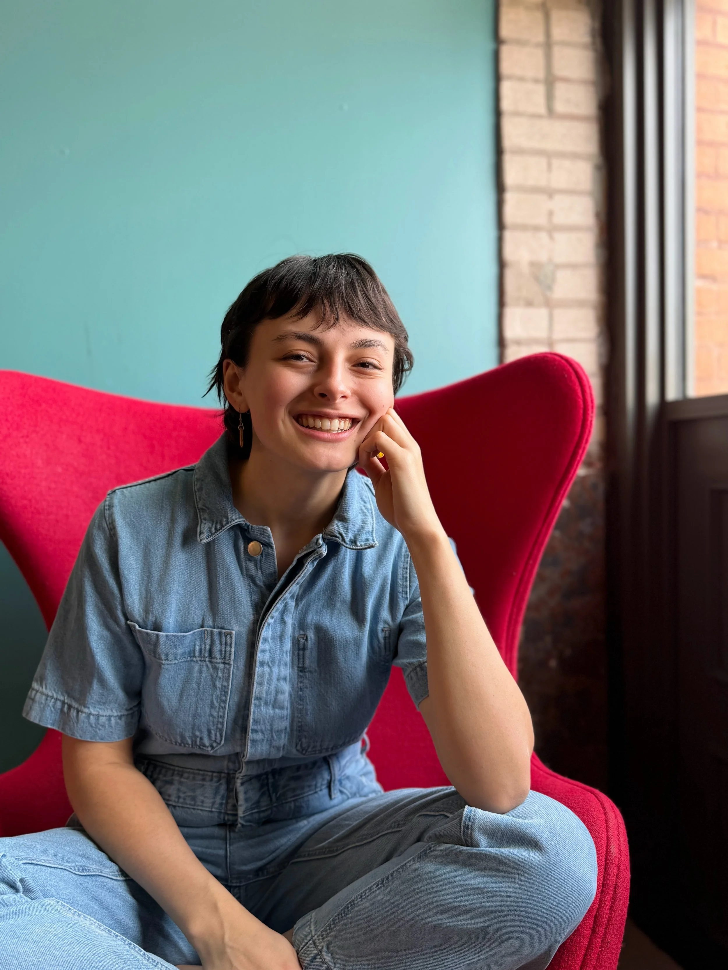 A woman with short dark hair and a denim outfit sitting on a red chair, smiling and resting her face on her hand, in a room with a teal wall and exposed brick near a window.