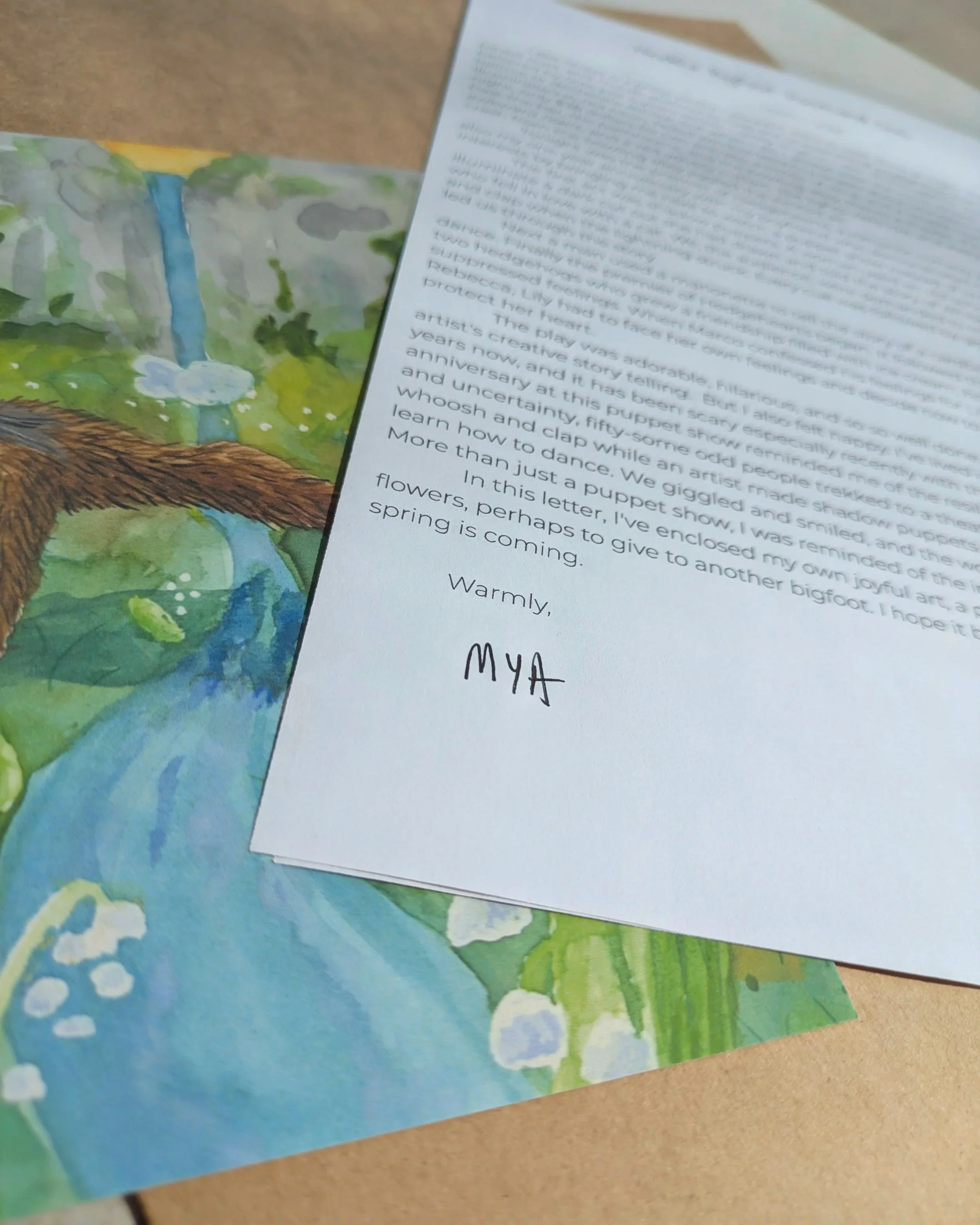 Close-up of a handwritten letter signed MYA, placed on a colorful watercolor painting of a landscape with a waterfall and greenery, on a wooden surface.