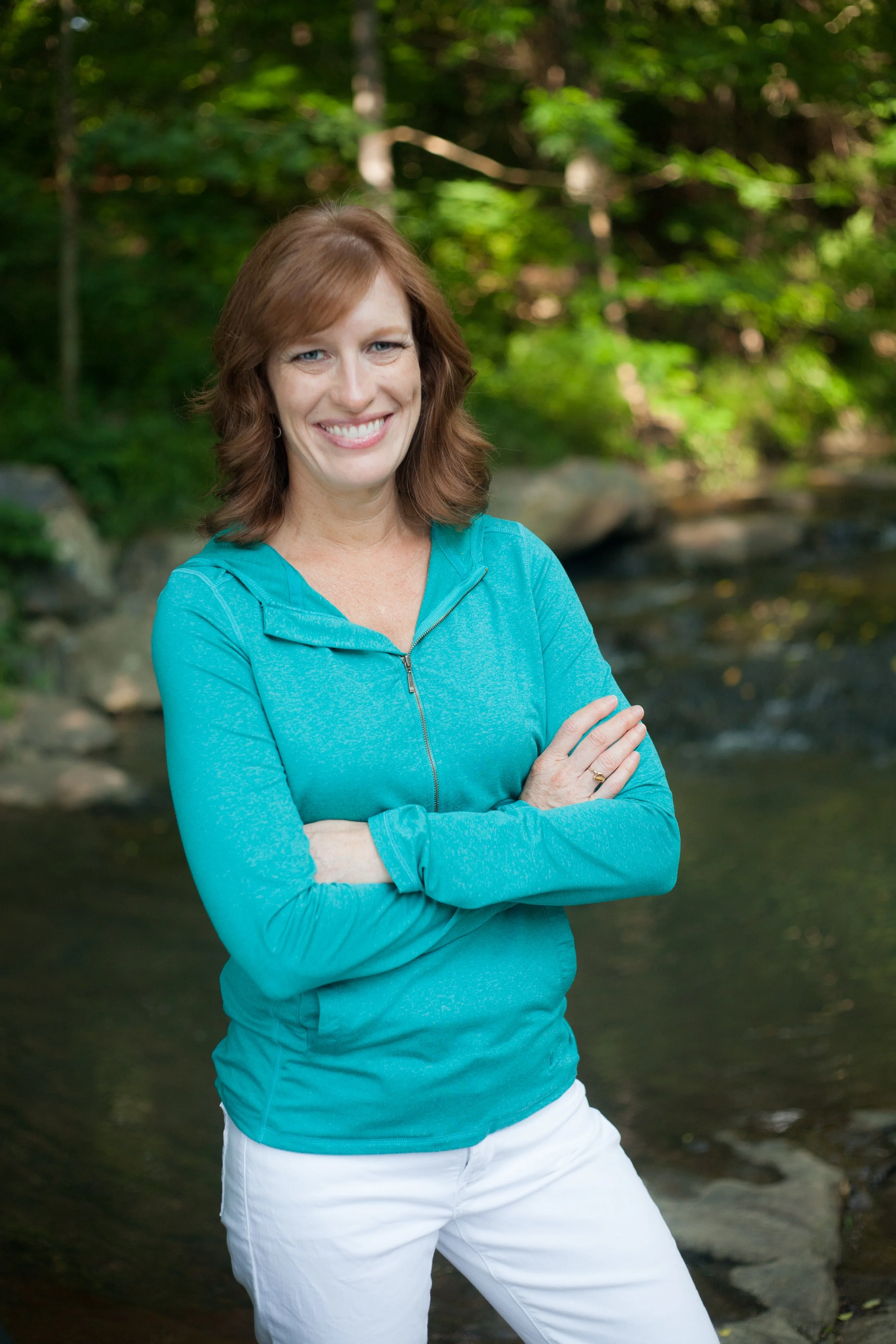 A smiling woman with shoulder-length red hair, wearing a turquoise zip-up jacket and white pants, standing outdoors next to a creek with green trees in the background.