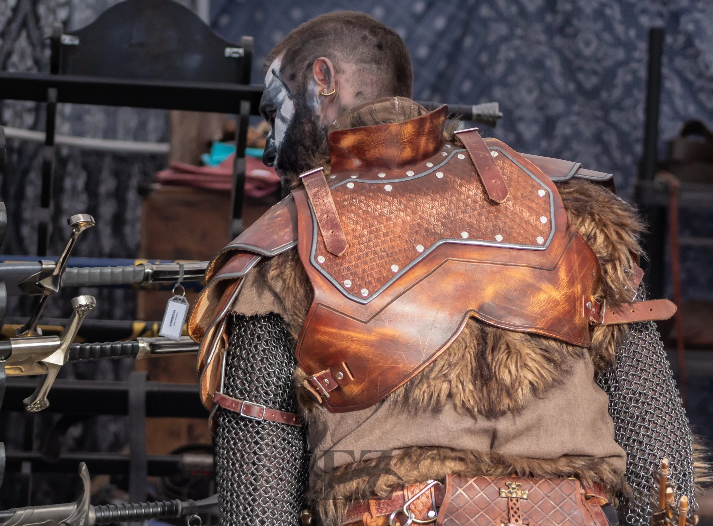 A person dressed as a medieval warrior, wearing chainmail, a leather chest armor, and fur accents, standing near swords displayed on racks.