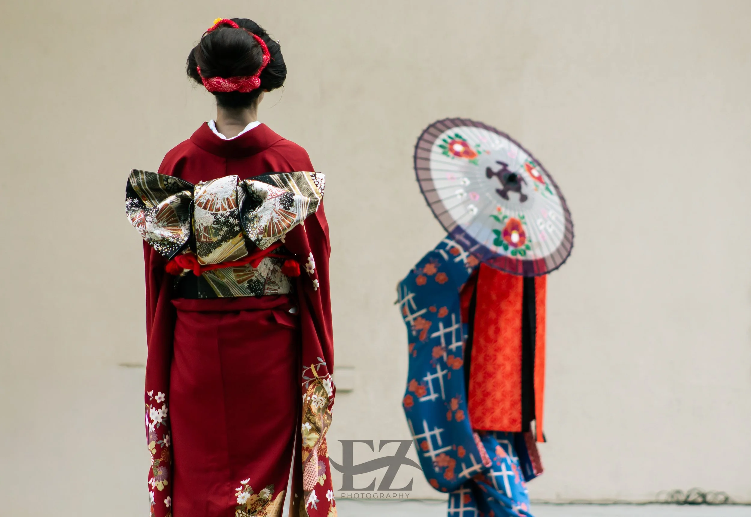 Two women dressed in traditional Japanese kimonos, one in a red kimono with an intricate bow-shaped obi, and the other wearing a blue and red kimono with a parasol.