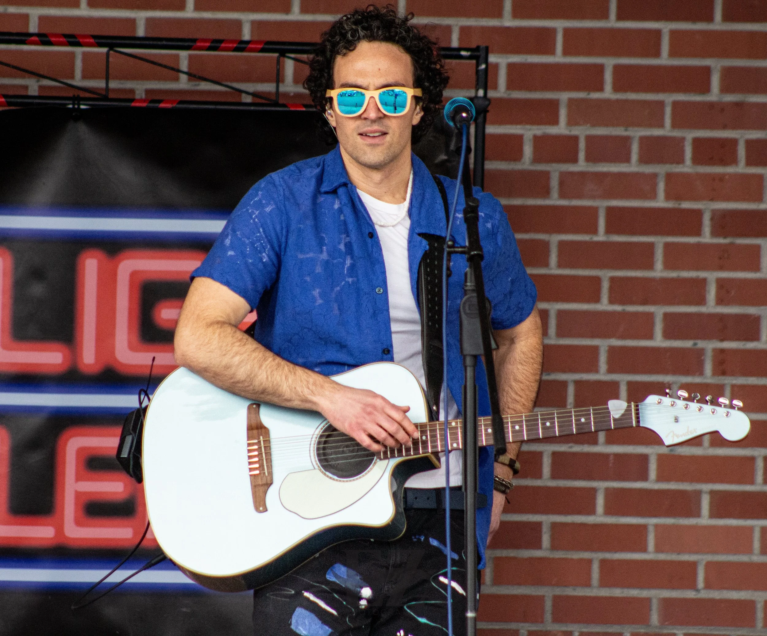 A man with curly hair and sunglasses wearing a blue shirt, holding an acoustic guitar, standing in front of a brick wall with a microphone.
