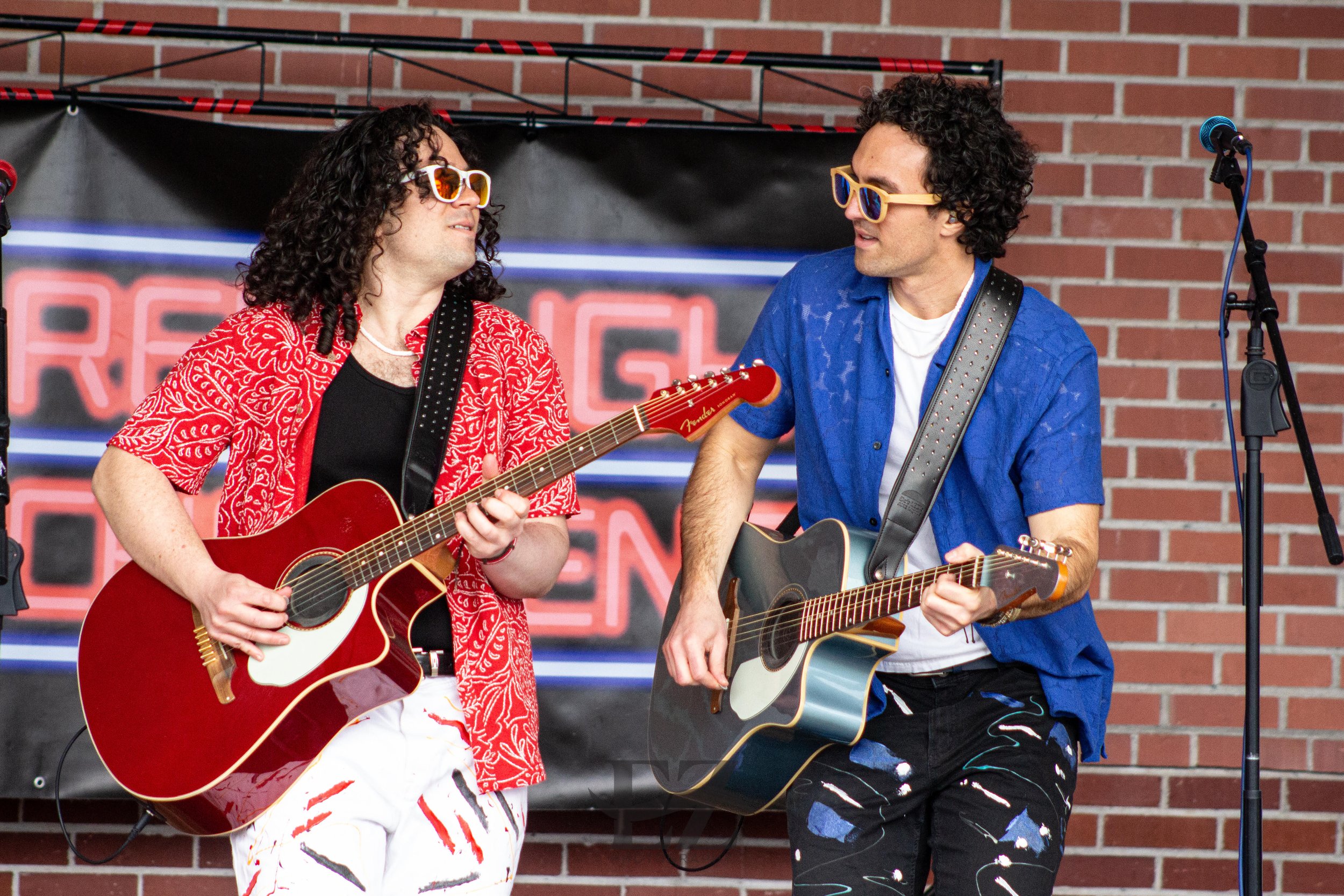 Two men playing guitars on stage with a brick wall background.