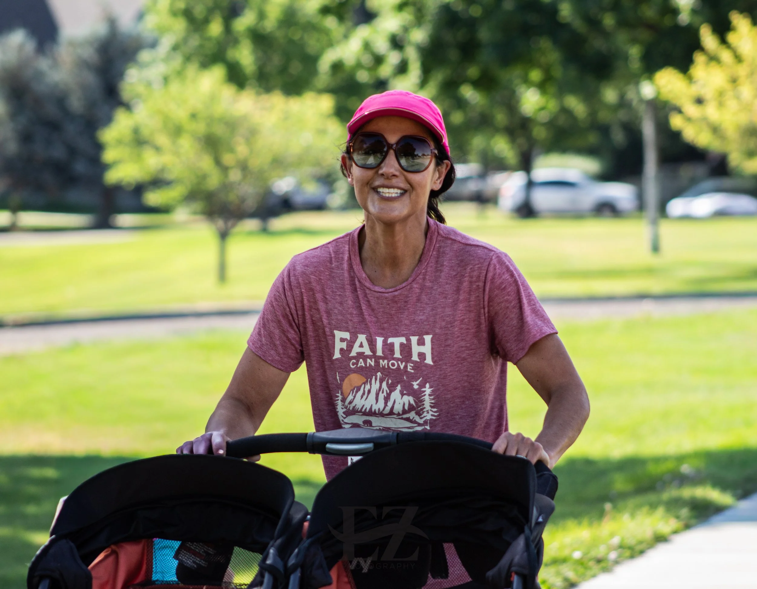 A woman in sunglasses and a pink cap smiling while walking outdoors with a stroller in a park on a sunny day.
