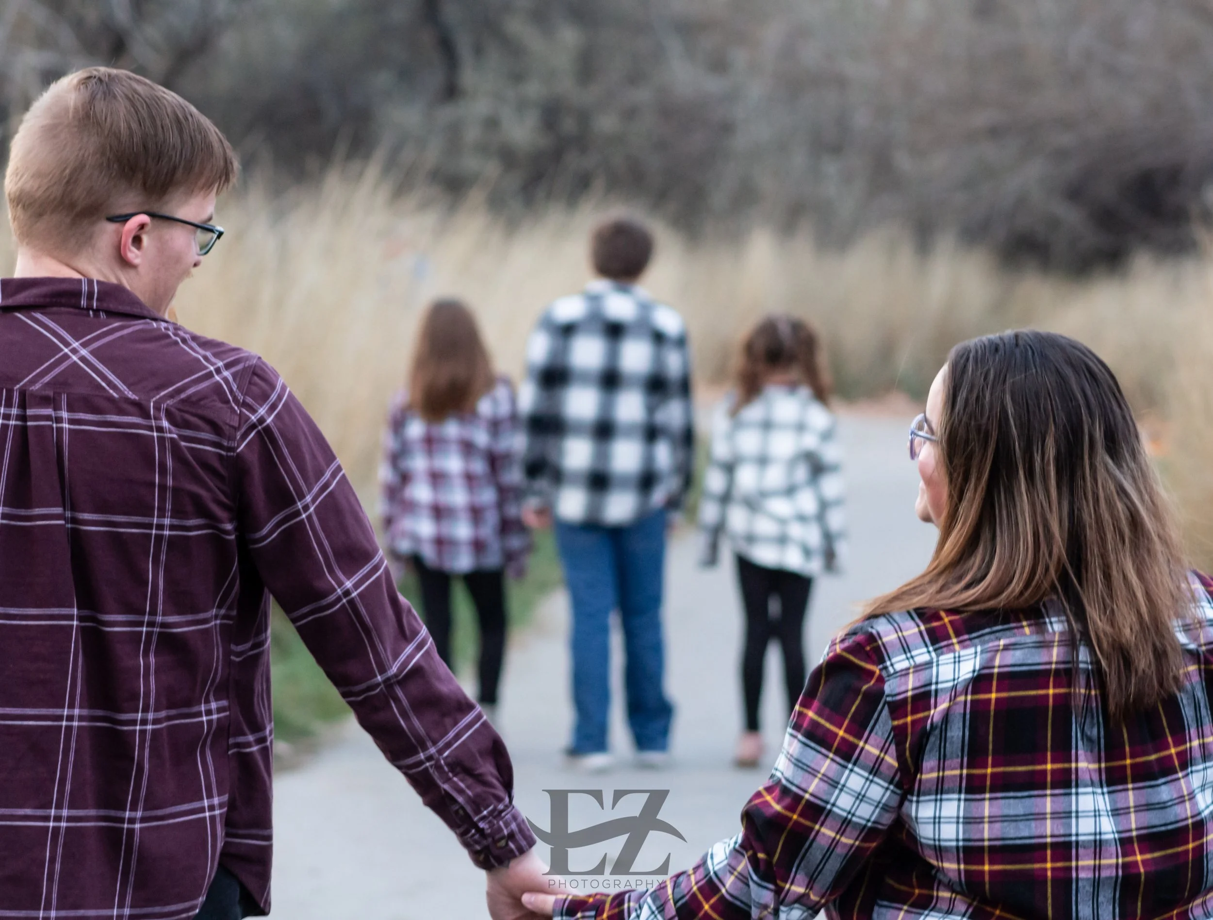 A couple holding hands walking on a trail in nature, with three children walking ahead of them, all wearing plaid shirts, in a scenic outdoor setting with tall grass and trees in the background.