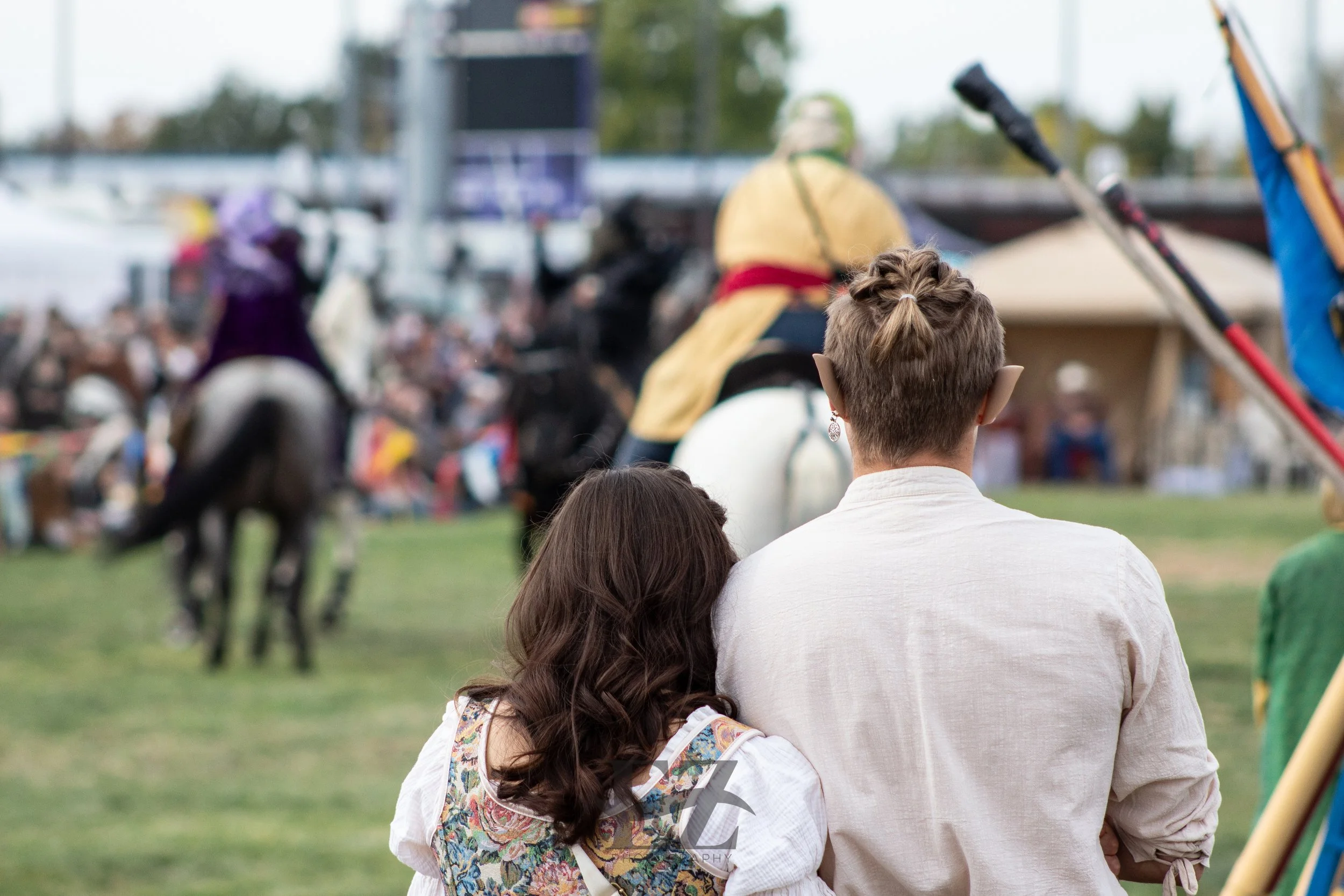 A couple watching a medieval-themed jousting event with people on horseback in historical costumes, on a grassy field, with a crowd in the background.