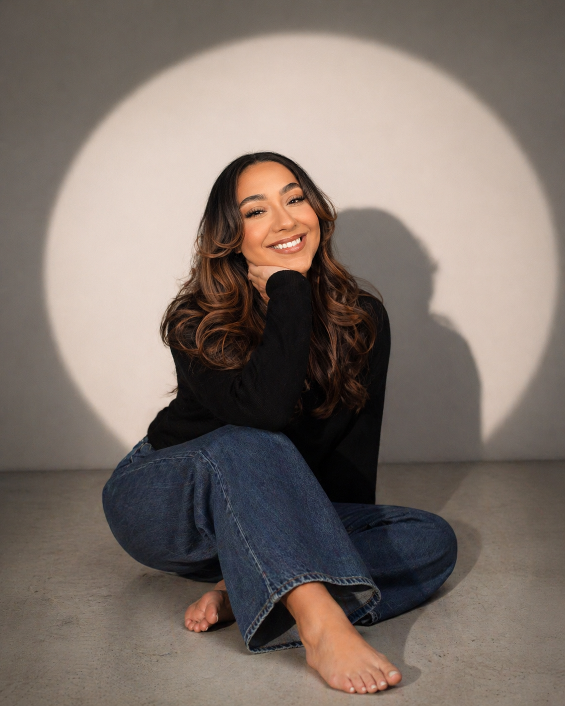 A smiling woman with long wavy hair sitting barefoot on the floor, wearing a black top and blue jeans, with a spotlight creating a circular shadow on the wall behind her.
