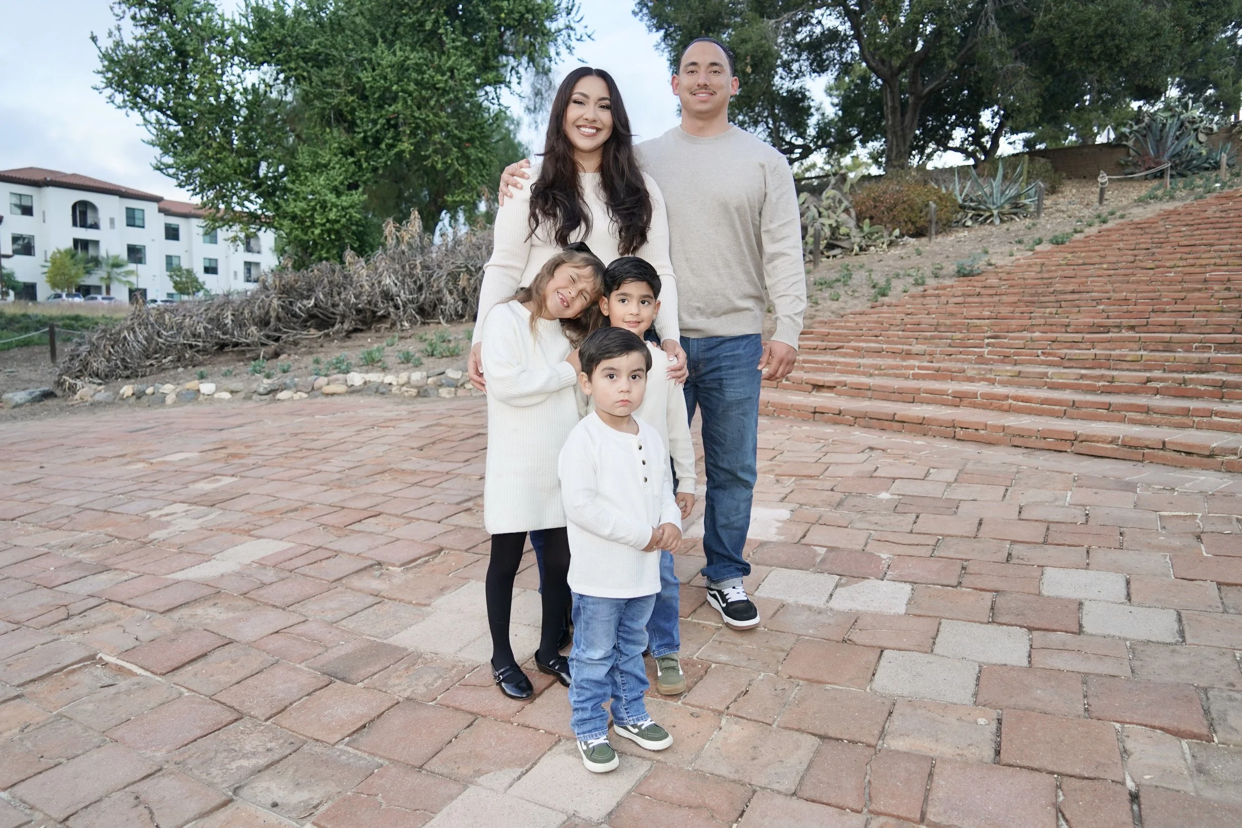 A family of five standing on a brick pathway outdoors, smiling at the camera. The background includes trees, succulents, and residential buildings.