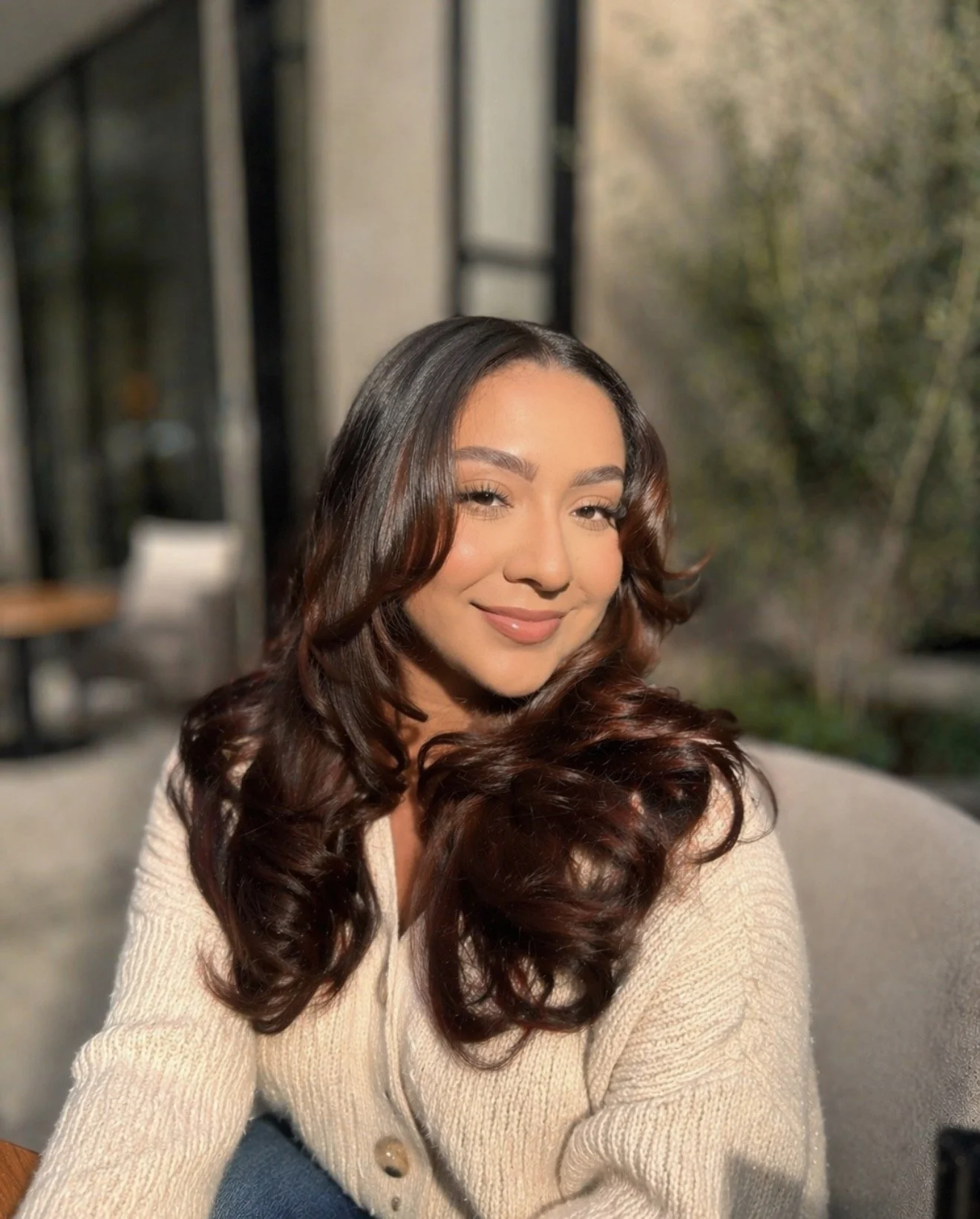 A woman with long, curly brown hair smiling and posing for a photo outside with blurred greenery and a building in the background.