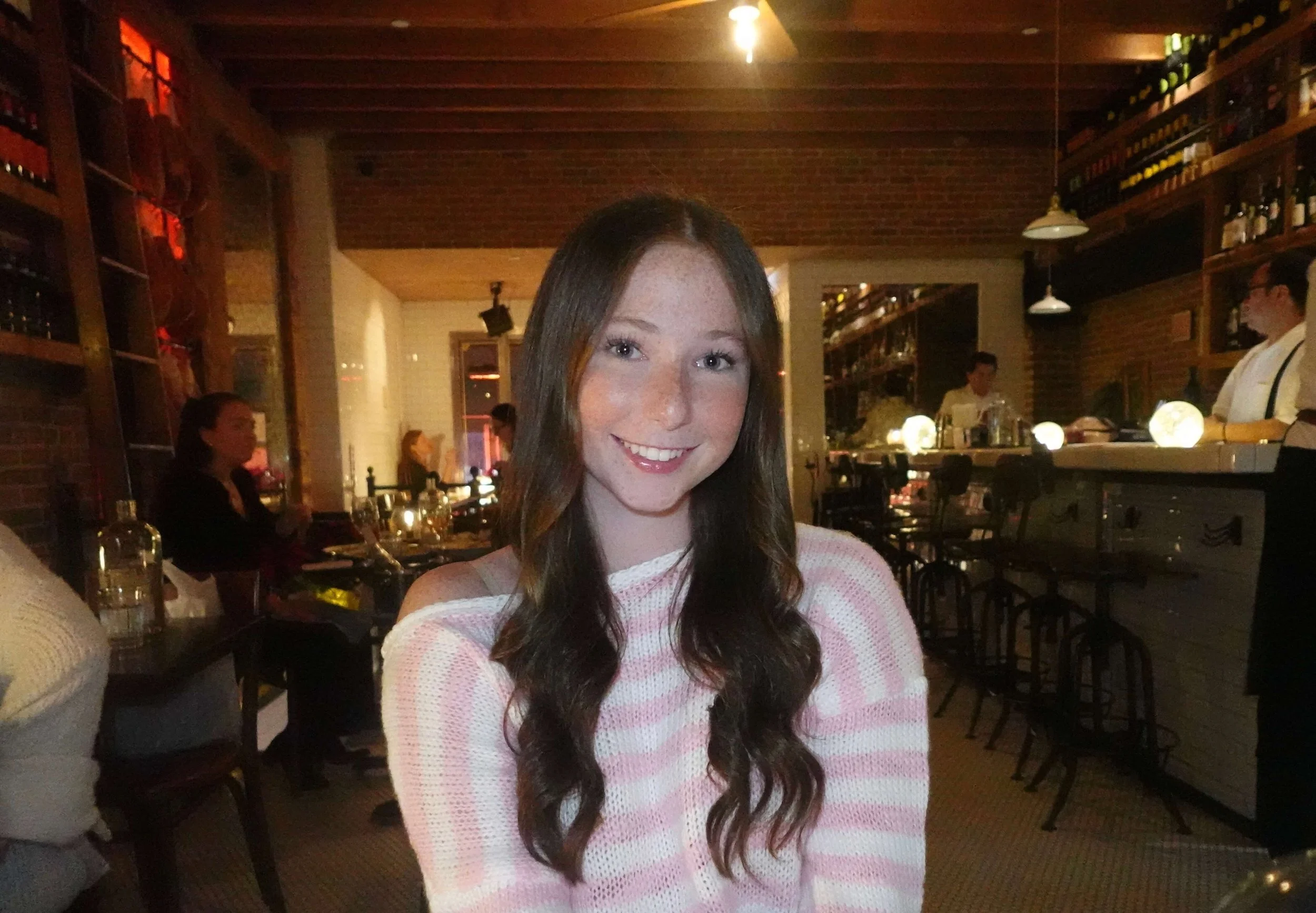 A young woman with long, wavy brown hair and fair skin is smiling at a restaurant or bar with warm lighting. She is wearing a white and pink striped sweater. In the background, there are shelves with bottles, and other patrons and staff can be seen.