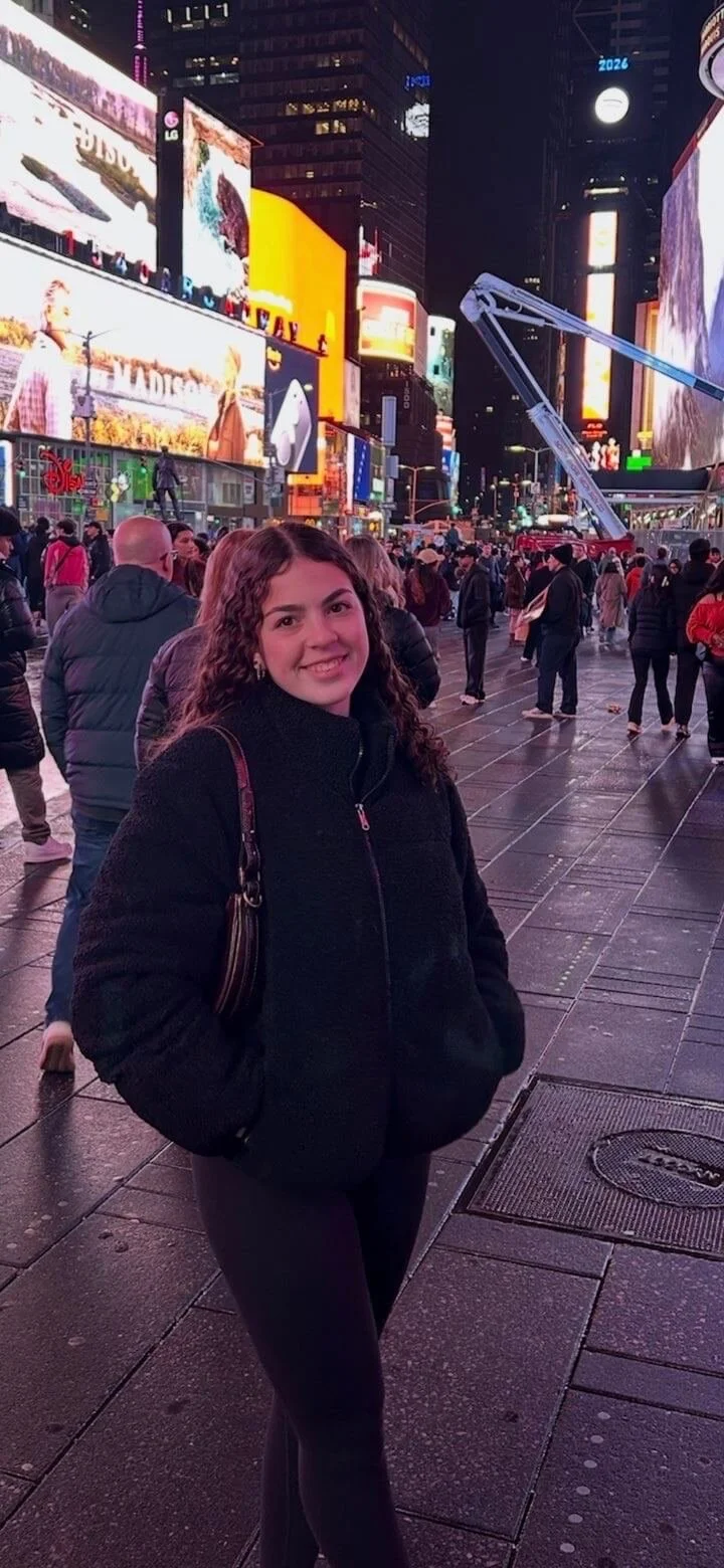 A young woman standing and smiling in Times Square at night, surrounded by bright digital billboards and a crowd of people.
