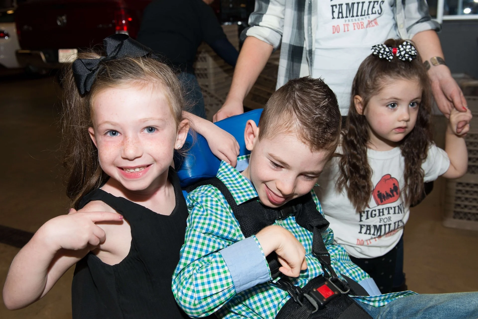 Four children sitting closely together, with one girl smiling and making a peace sign, a boy looking down at his lap, and two girls with long hair, one wearing a camouflage shirt and the other wearing a white shirt that says "Fighting for Families," all with bows in their hair, in what appears to be a busy indoor setting.