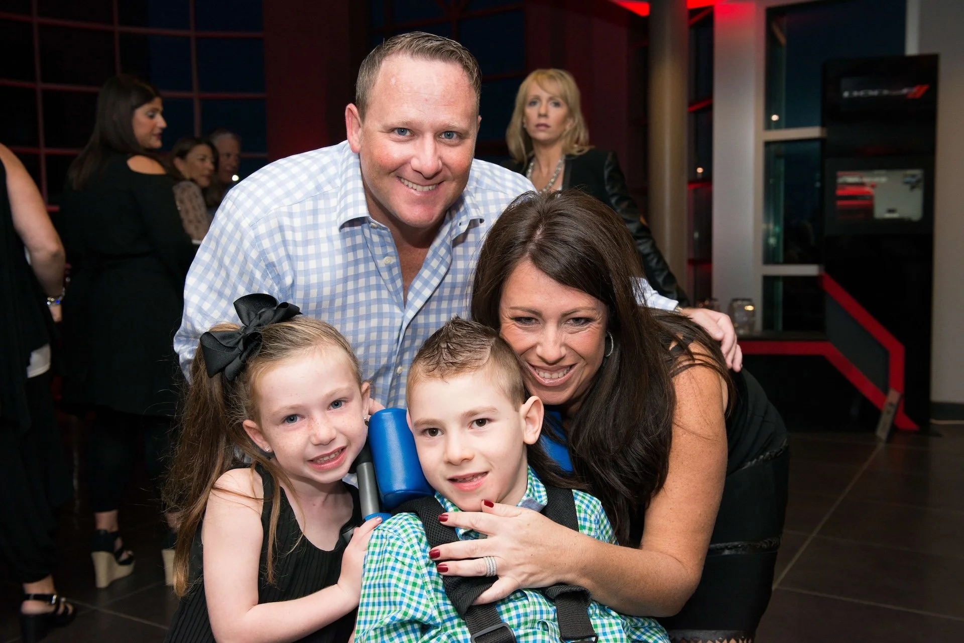Group of four people, including two children, hugging and smiling at a social event. Two adults and two children in the foreground, with other guests in the background.
