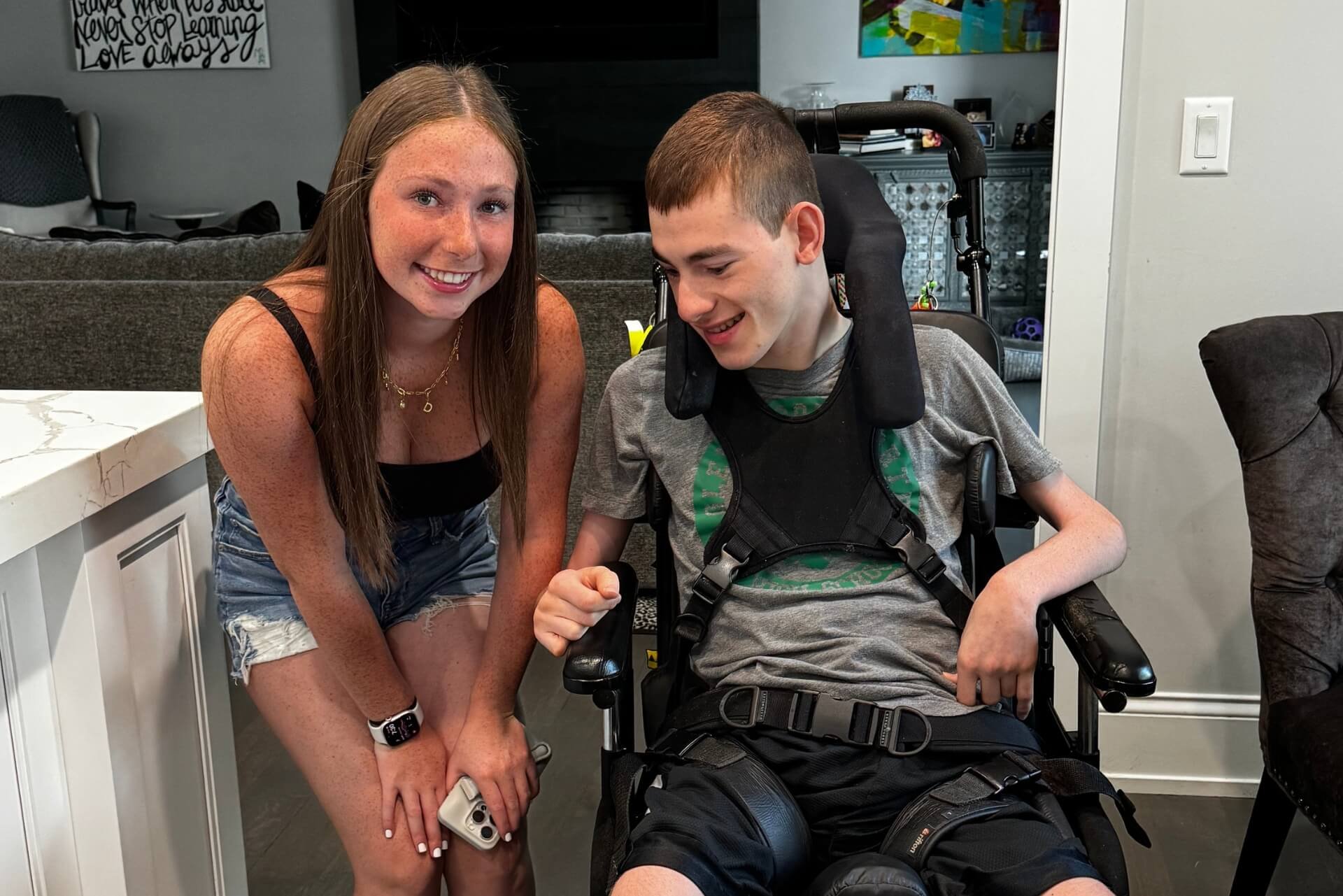 A smiling woman with long brown hair and a necklace kneels beside a young man in a motorized wheelchair. The woman is holding a smartphone in her hand.